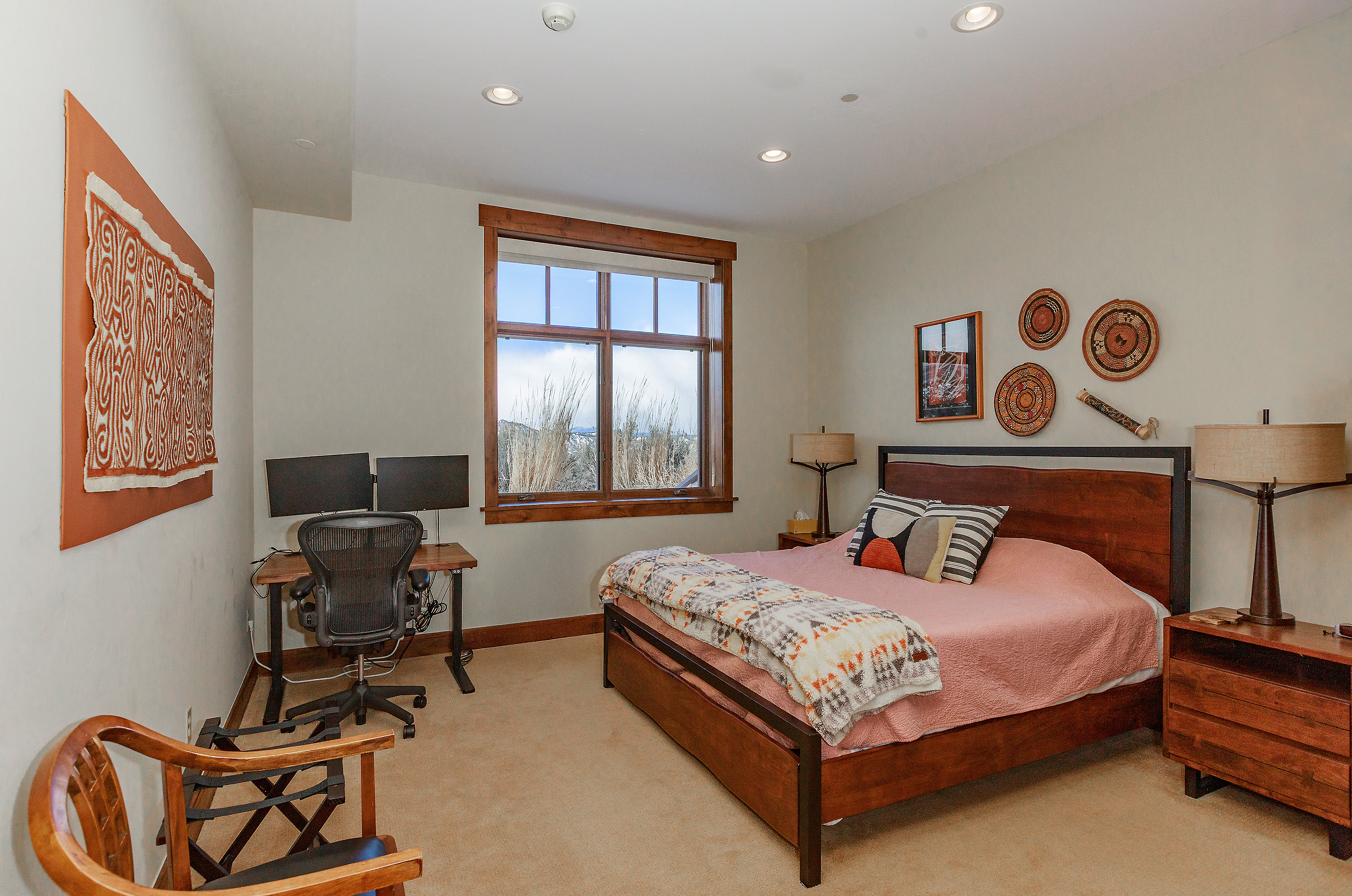This primary bedroom features a warm, rustic aesthetic with a wooden bed frame, matching nightstands, and a desk area positioned by a large window that offers a scenic view. The room is decorated with Southwestern-inspired wall art and textiles, creating a cozy and inviting atmosphere. The perspective is a wide-angle shot from the corner of the room, capturing the functional layout that blends sleeping and workspace areas.