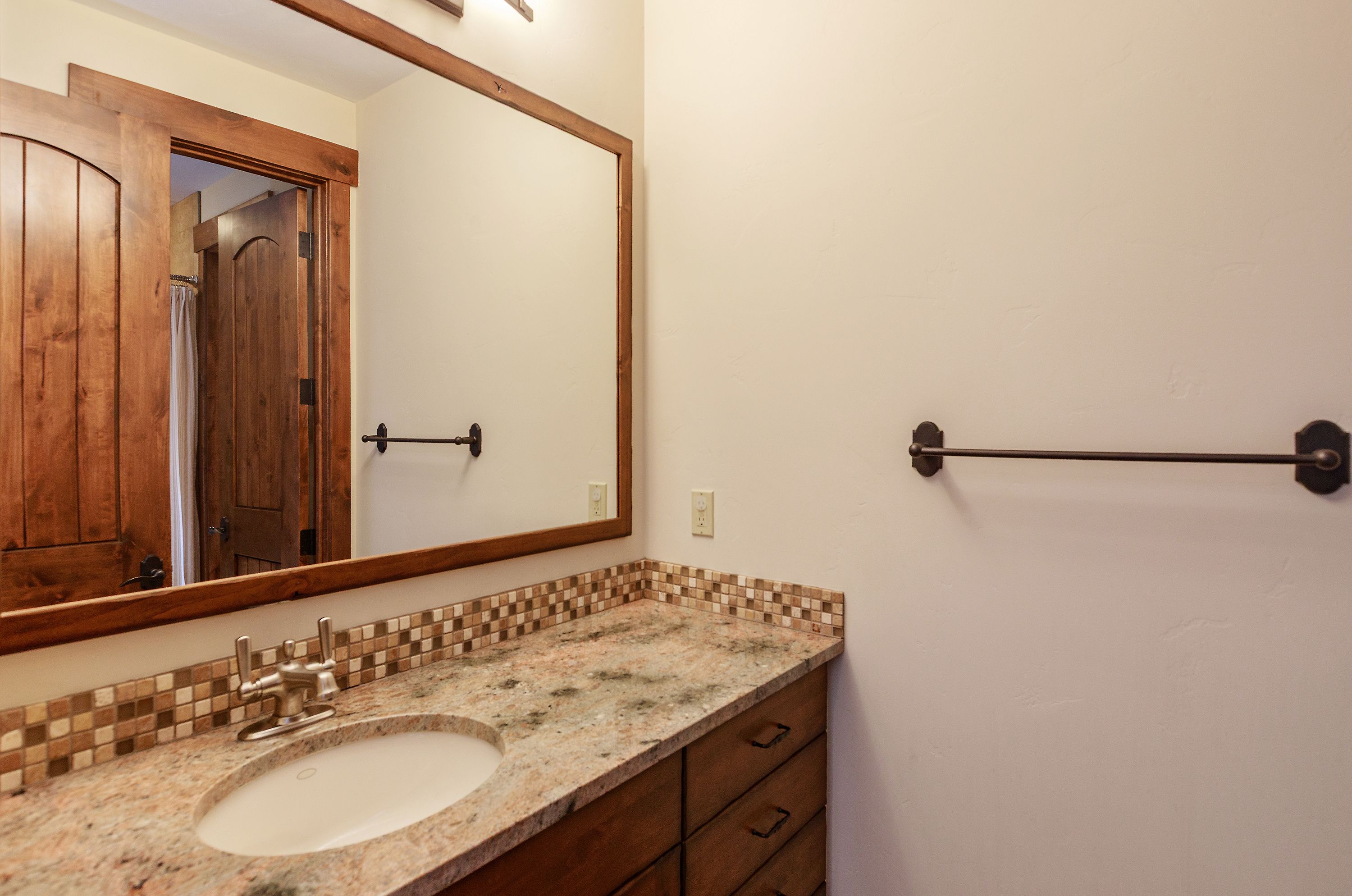 This bathroom features a rustic-inspired vanity with a granite countertop, an undermount sink, and a decorative mosaic tile backsplash. A large, wood-framed mirror hangs above the vanity, reflecting the adjacent doorway, while a dark metal towel bar is mounted on the plain wall to the right. The space is characterized by warm, earthy tones and a clean, functional layout.