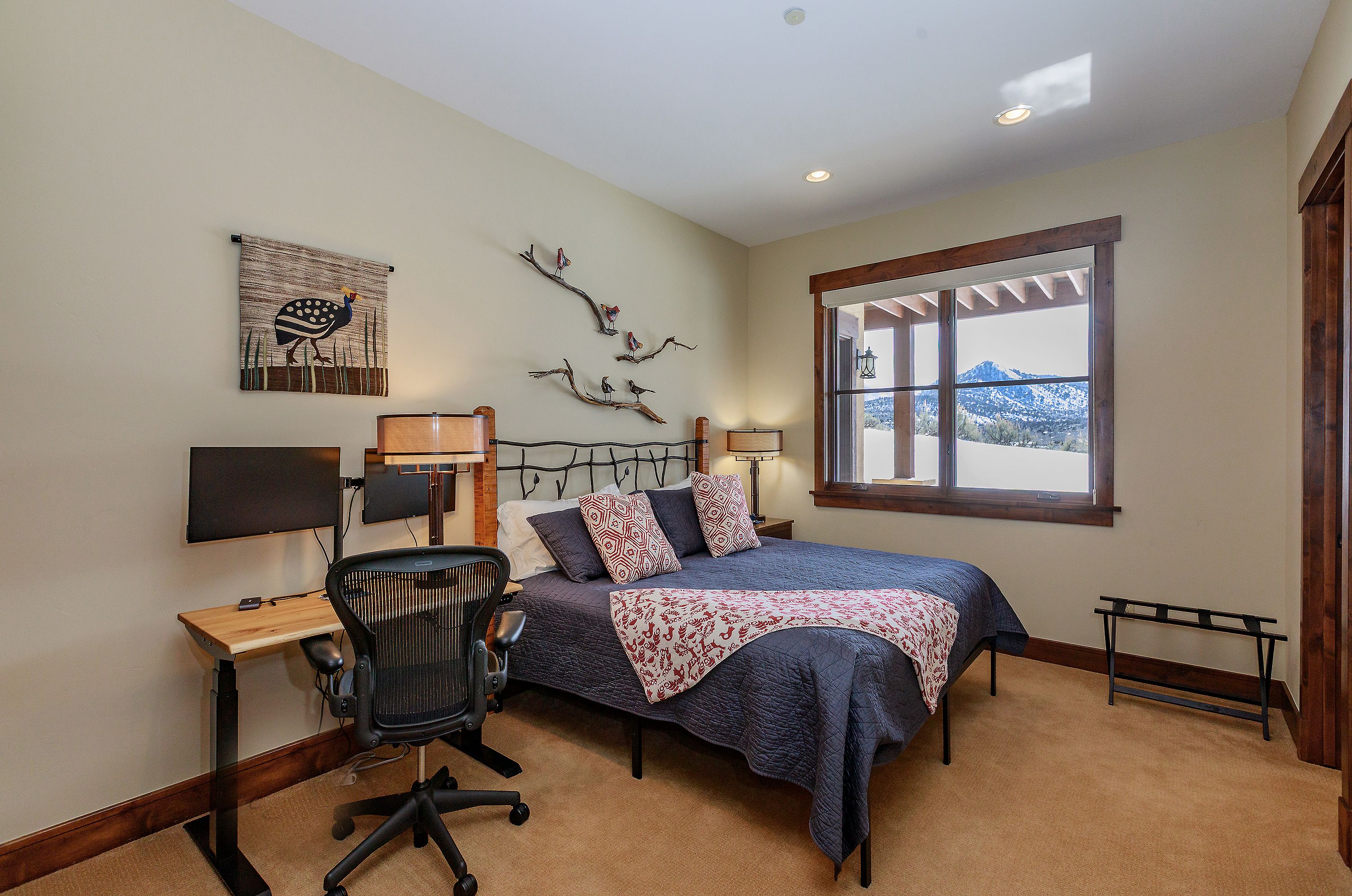 This cozy bedroom features a rustic aesthetic with a dark metal bed frame, a patterned quilt, and decorative branch wall art. A functional workspace is integrated into the room with a modern ergonomic chair and a standing desk, while a large window provides a stunning view of snow-covered mountains. The warm lighting and neutral color palette create a comfortable and inviting atmosphere.