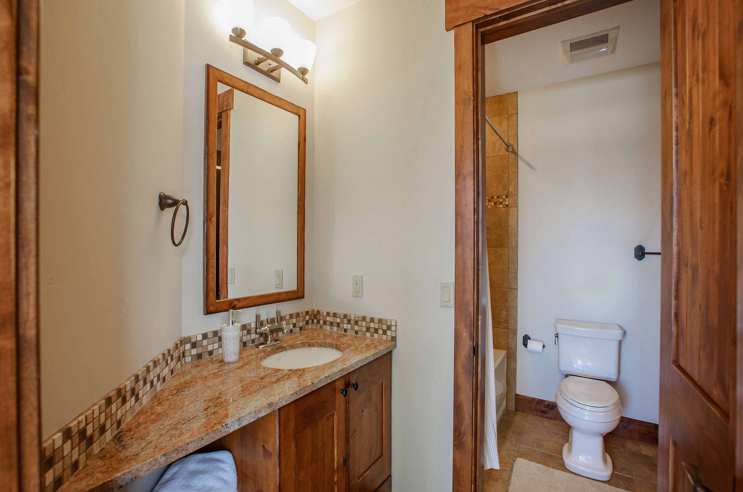 This bathroom features a rustic aesthetic with a warm wood vanity, granite countertop, and a decorative mosaic tile backsplash. The layout includes a separate water closet area with a toilet and shower, framed by a sturdy wooden doorway that adds to the cabin-like charm. The perspective is a straight-on shot from the vanity area, highlighting the functional flow and cozy, earthy color palette.