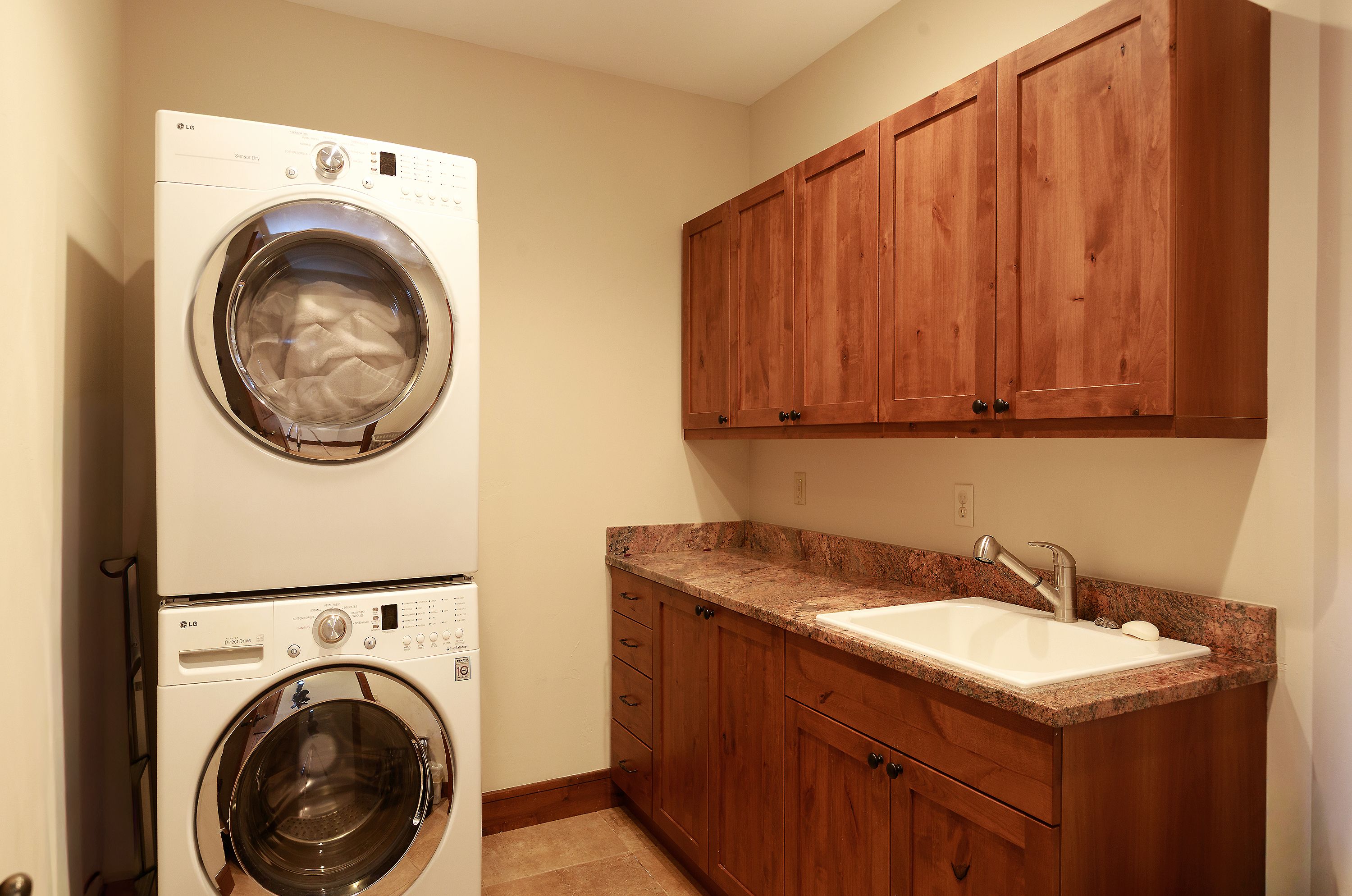 This functional laundry room features a stacked LG washer and dryer unit positioned next to a custom wood cabinetry setup with a granite countertop and an integrated utility sink. The warm-toned wood cabinets and neutral wall color create a cohesive and practical workspace. The perspective is a straight-on eye-level shot, highlighting the efficient use of space and the clean, well-maintained condition of the room.