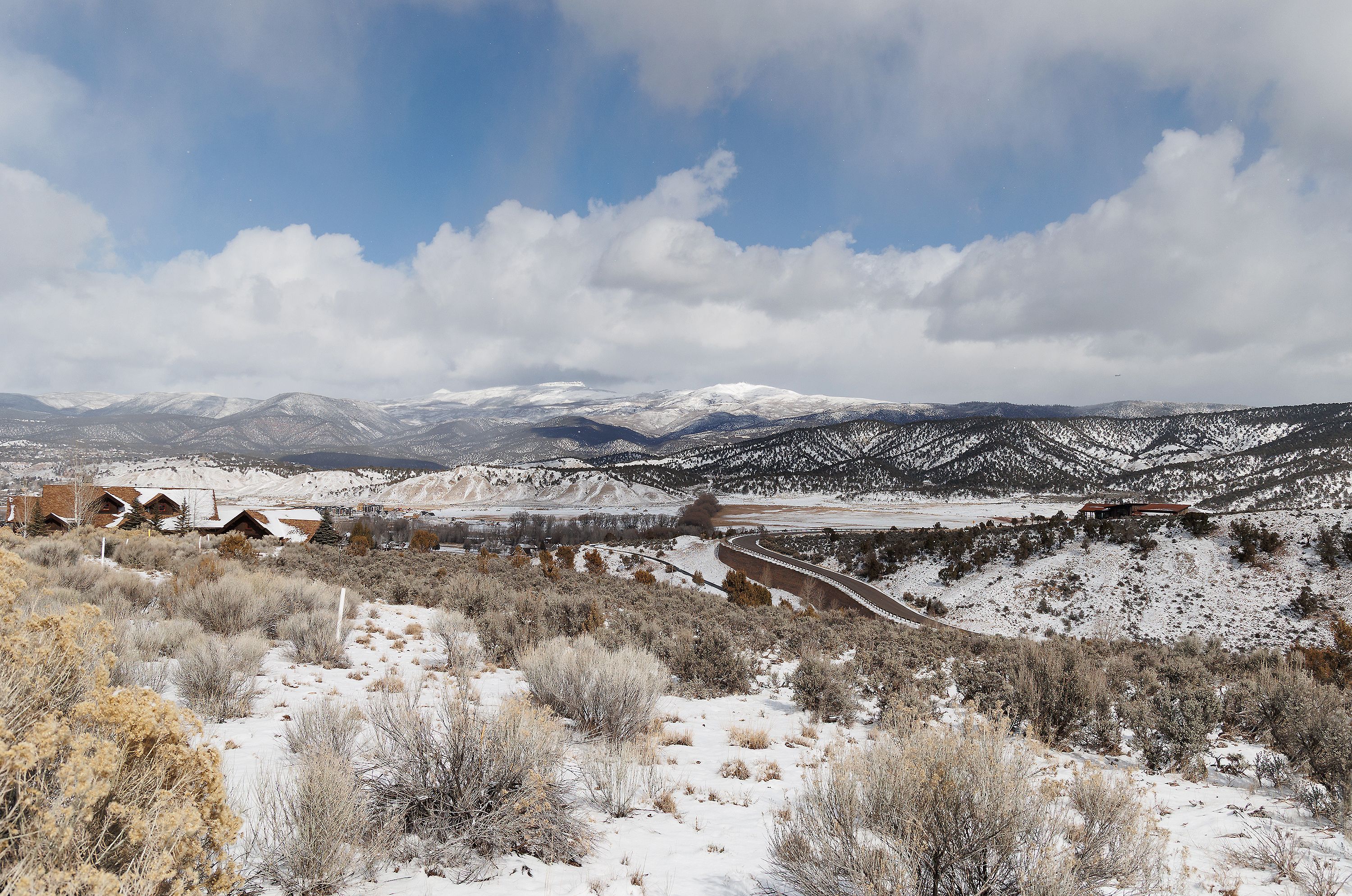 This wide-angle landscape shot captures a sprawling, snow-dusted mountain terrain under a dramatic, cloud-filled sky. The scene features a winding road cutting through the rugged, sagebrush-covered hills, with residential structures nestled into the landscape on the left and right. The composition emphasizes the vast, serene beauty of the high-altitude environment, highlighting the property's integration into the natural, mountainous surroundings.