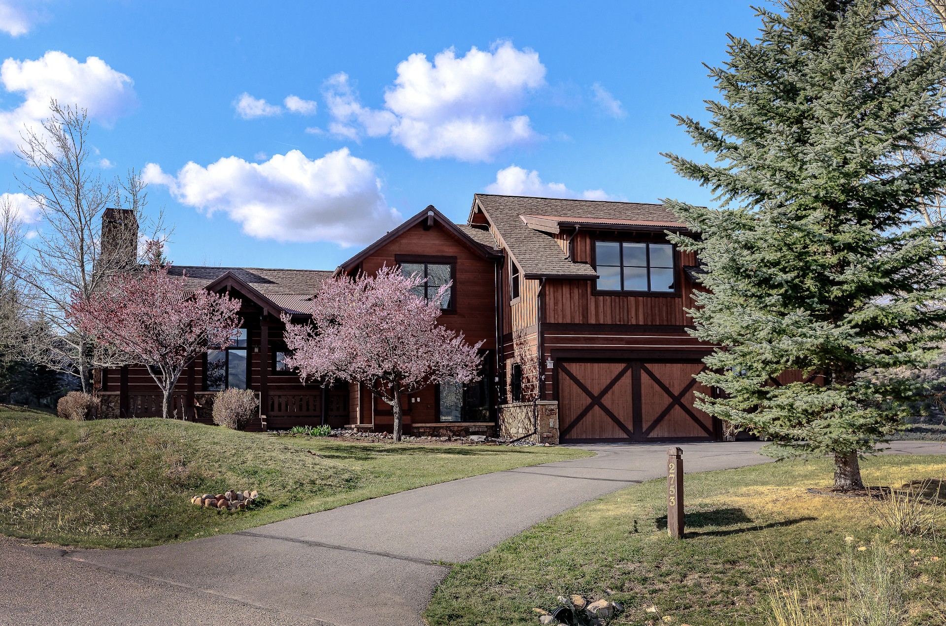 This stunning mountain-style residence features a warm, rustic aesthetic with wood siding, stone accents, and a multi-gabled roofline. The property is beautifully landscaped with vibrant pink flowering trees in the front yard, a paved driveway leading to a multi-car garage, and a large evergreen tree framing the right side. The scene is captured from a low-angle, eye-level perspective on a bright, sunny day, emphasizing the home's inviting curb appeal and natural surroundings.
