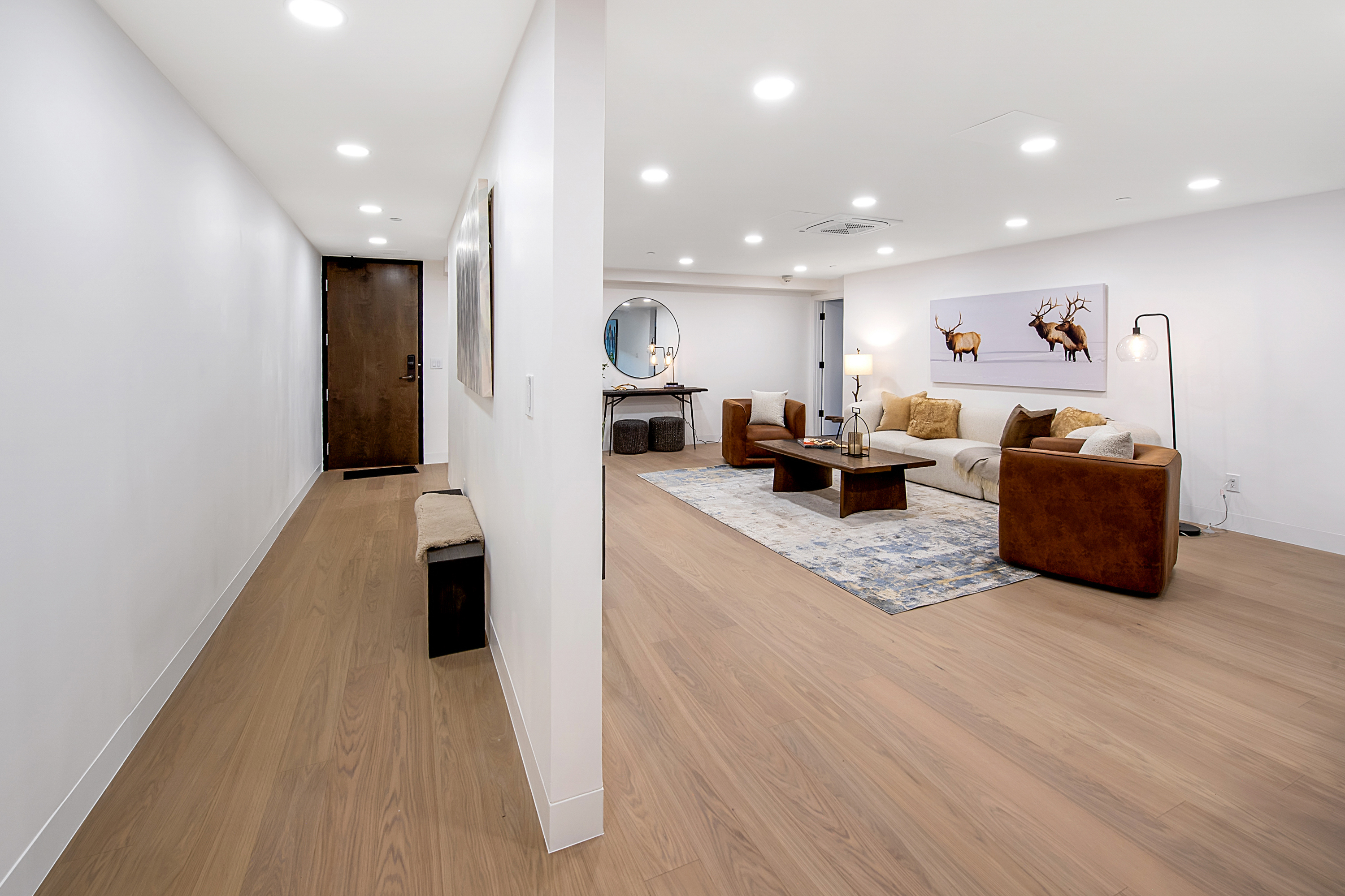This spacious, open-concept living area features light wood flooring and a neutral color palette, creating a bright and inviting atmosphere. The room is anchored by a large, plush sofa, two leather armchairs, and a modern coffee table, all set upon a textured area rug beneath a striking elk-themed wall art piece. A hallway leads to the front entrance, showcasing a clean, contemporary design that seamlessly blends functionality with sophisticated comfort.