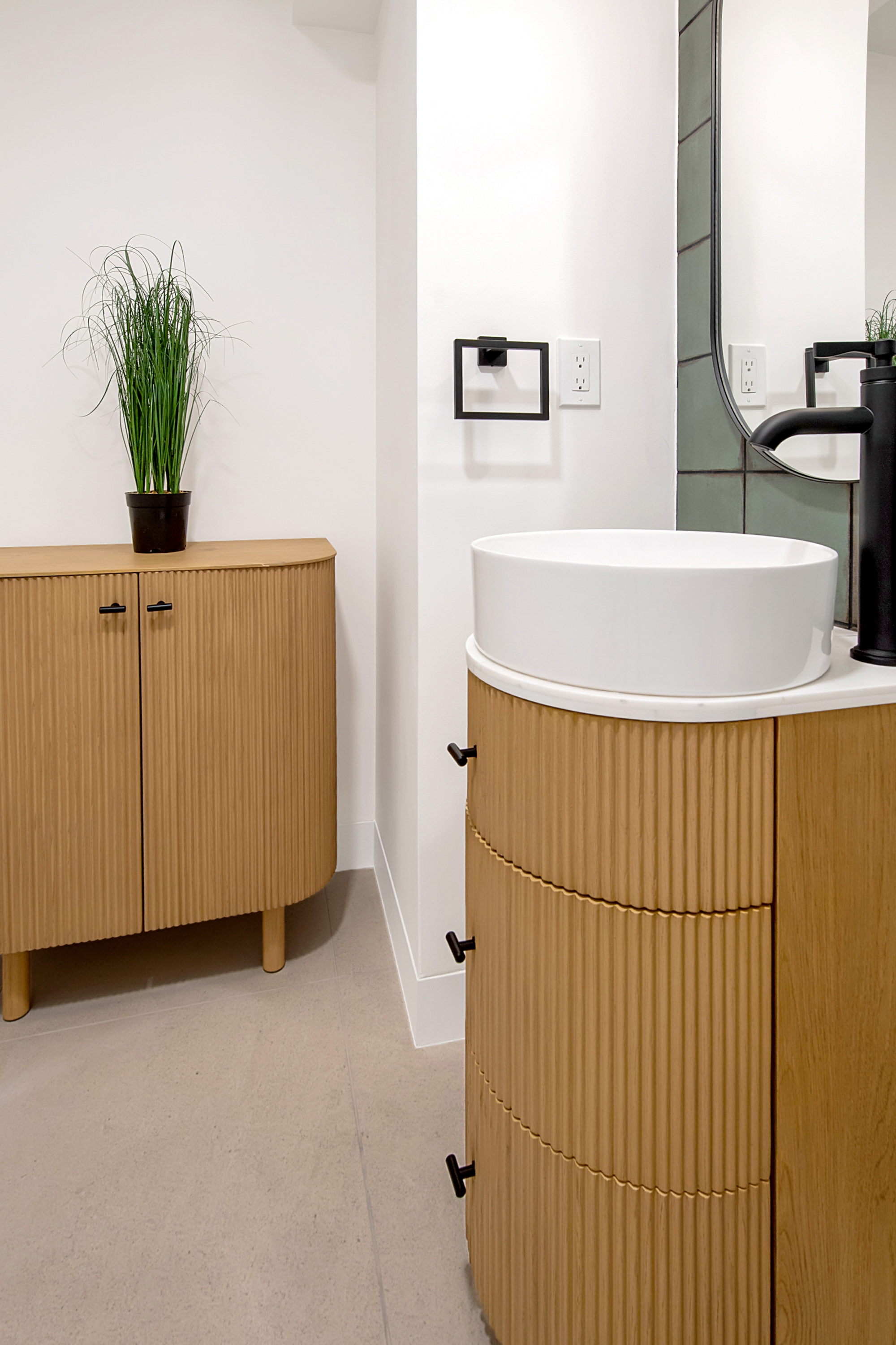 This modern bathroom features a stylish, curved vanity with a fluted wood finish and a white vessel sink, complemented by a matching fluted storage cabinet nearby. The space is accented with matte black hardware, a sleek mirror, and sage green wall tiles, creating a sophisticated and contemporary aesthetic. The perspective is a close-up, eye-level shot that highlights the high-end textures and clean lines of the fixtures.