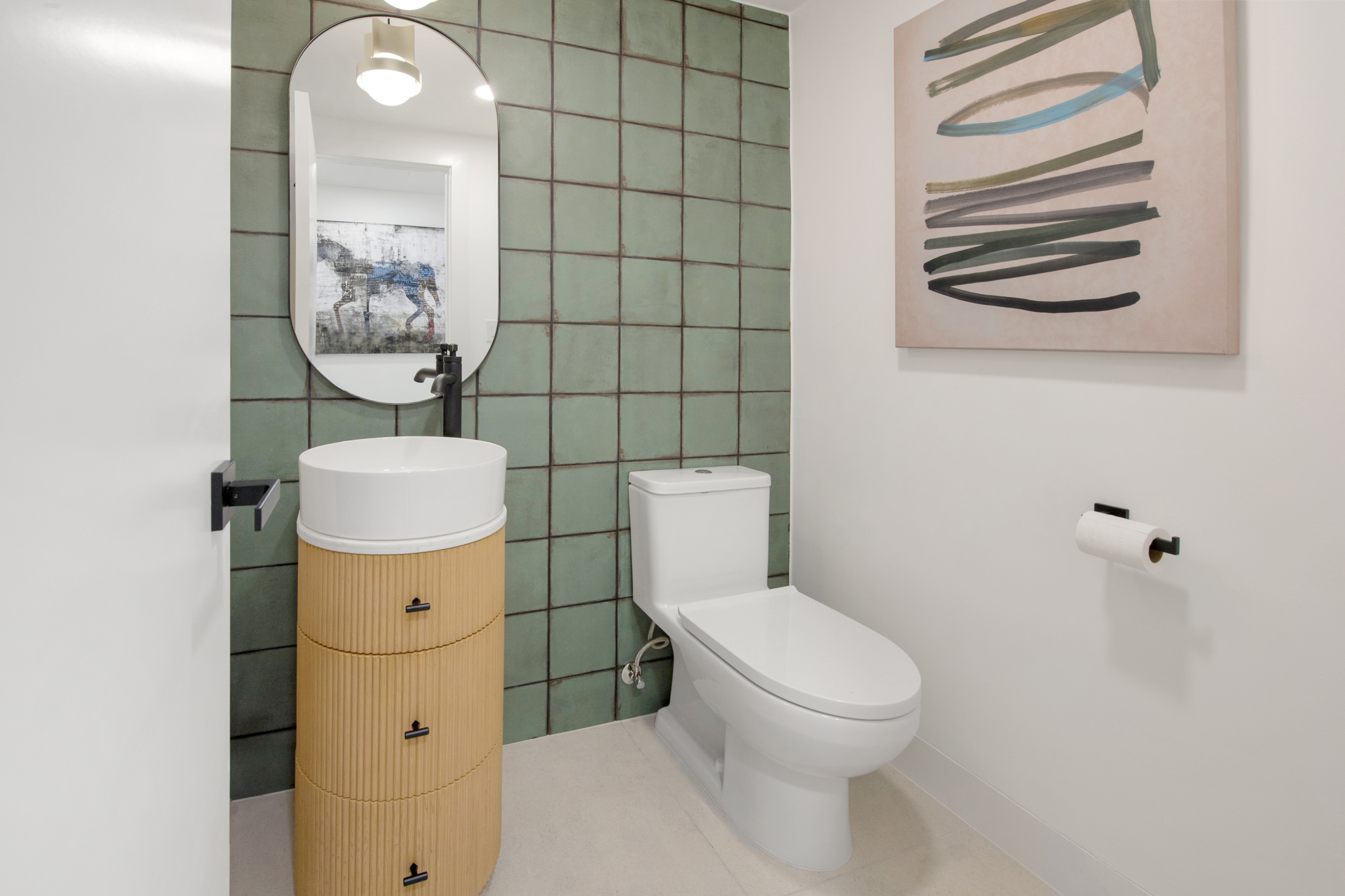 This modern powder room features a striking green square-tiled accent wall that serves as a focal point behind a sleek, oval mirror and a unique, cylindrical fluted vanity. The space is completed by a white pedestal-style toilet and minimalist black hardware, creating a contemporary and artistic aesthetic. The composition is captured from a straight-on perspective, highlighting the clean lines and bold color palette of this compact bathroom.