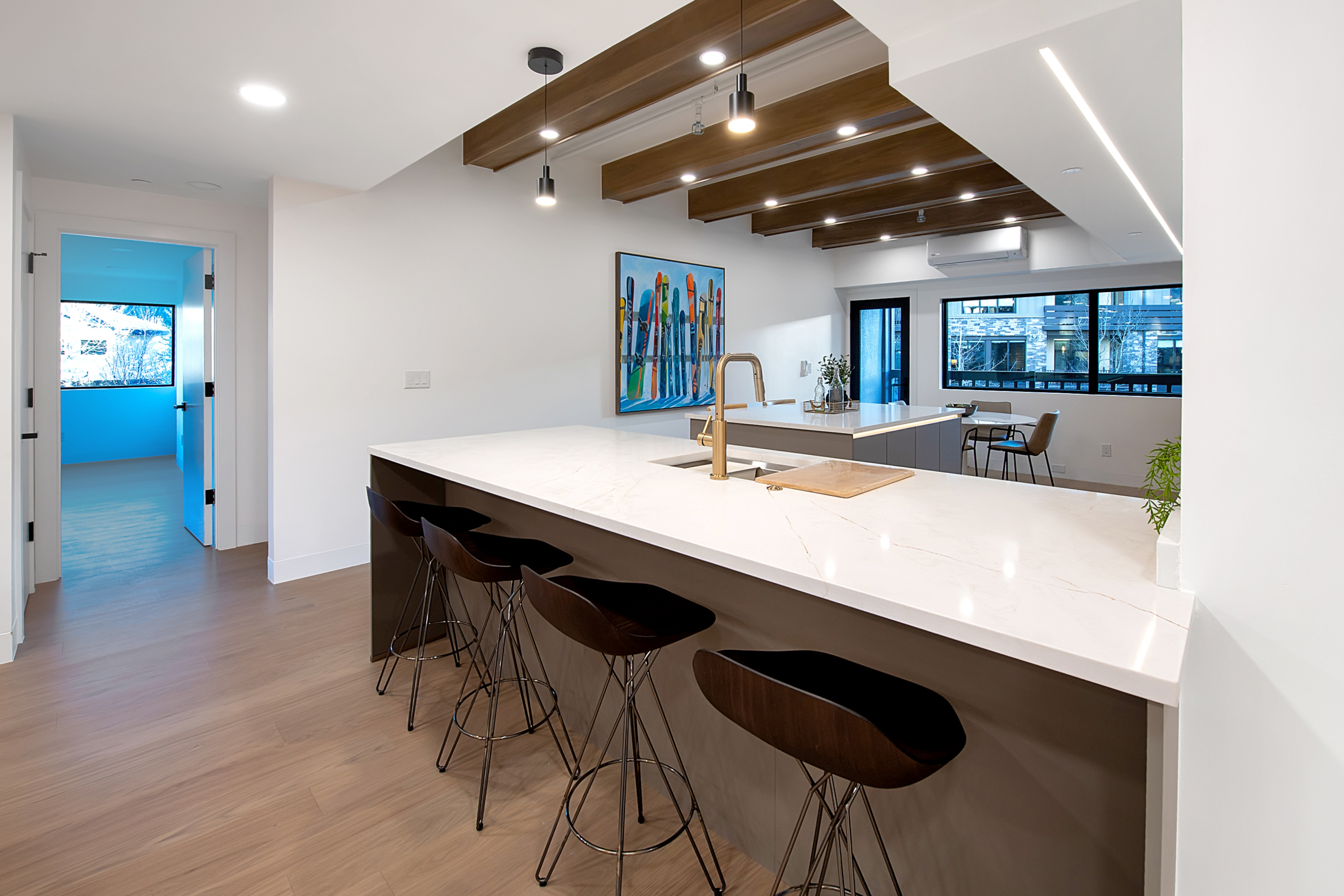 This modern, open-concept kitchen features a large island with a white quartz countertop and dark wood bar stools, perfect for casual dining. The space is highlighted by striking wooden ceiling beams, recessed lighting, and a vibrant piece of ski-themed wall art that adds a playful touch. The clean lines, neutral color palette, and seamless flow into the adjacent dining area create a sophisticated and inviting atmosphere.