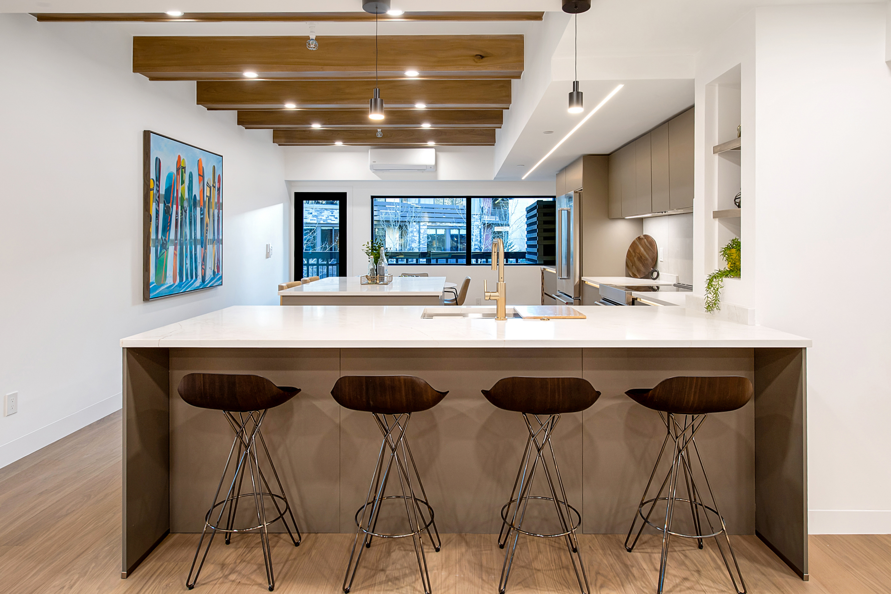 This modern, open-concept kitchen features a large central island with a white quartz countertop and four stylish mid-century modern bar stools. The space is defined by striking wooden ceiling beams, sleek taupe cabinetry, and a vibrant piece of ski-themed wall art that adds a playful touch. The perspective is a straight-on, eye-level shot that highlights the seamless flow between the kitchen, dining area, and the large window overlooking the exterior.