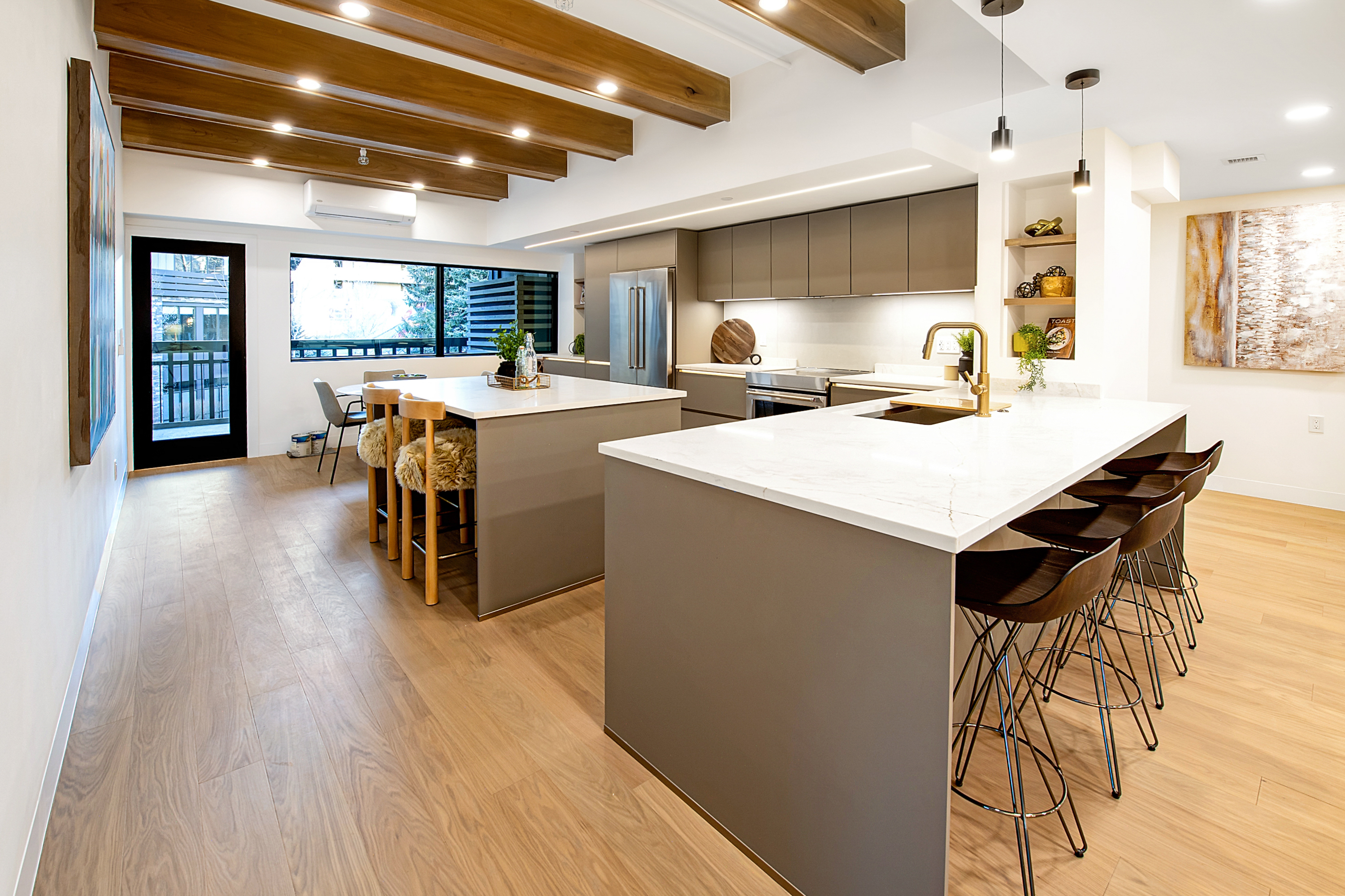 This modern, open-concept kitchen features dual islands with sleek grey cabinetry and white quartz countertops, complemented by warm wood-toned flooring and exposed ceiling beams. The space is illuminated by contemporary pendant lighting and recessed lights, creating a bright and inviting atmosphere. A large window provides a view of the exterior, while the clean lines and minimalist aesthetic offer a sophisticated, high-end feel.