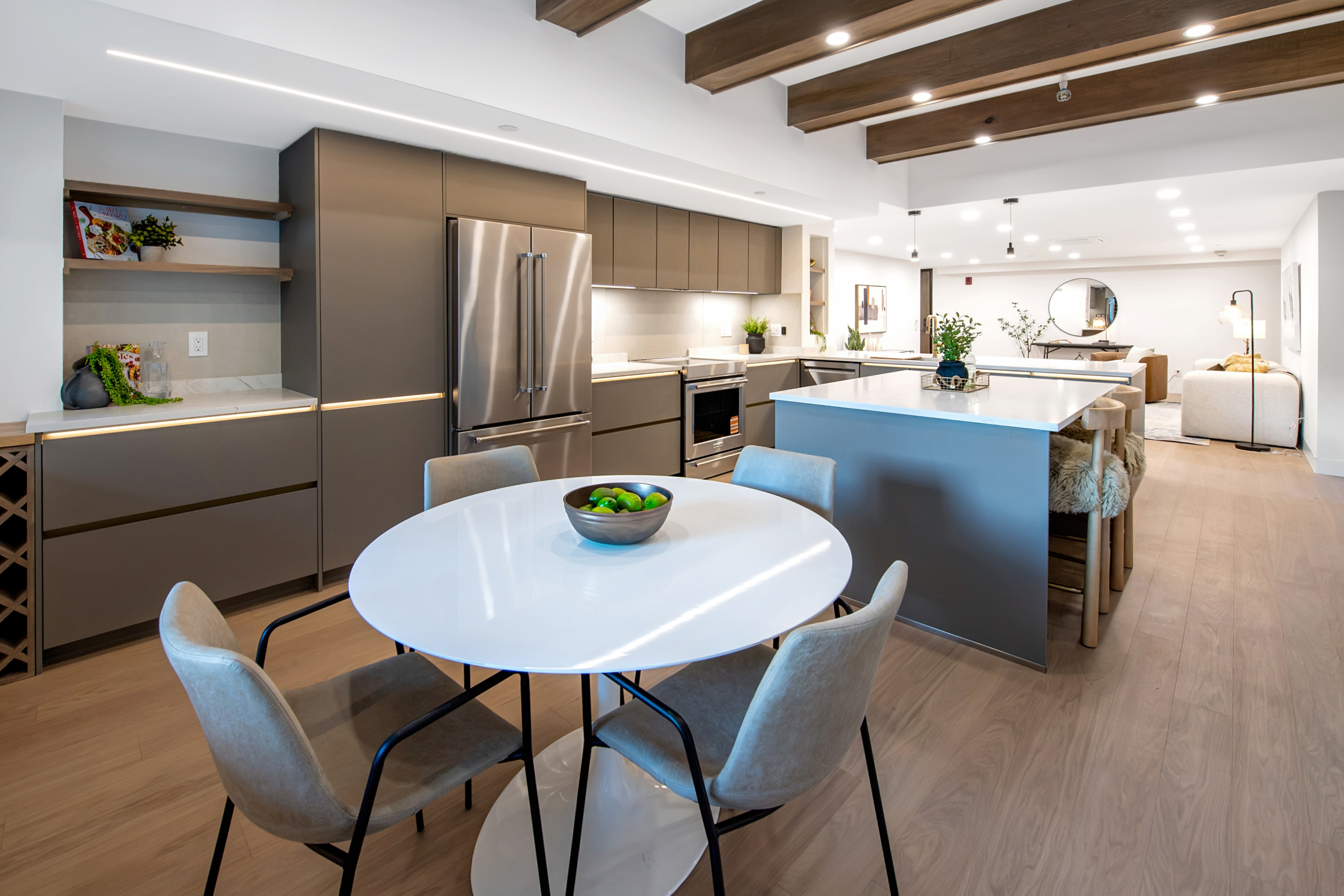 This modern, open-concept kitchen features sleek matte-grey cabinetry, stainless steel appliances, and a central island with a white countertop and bar seating. A round dining table sits in the foreground, while exposed wooden ceiling beams add warmth to the contemporary design. The space flows seamlessly into a bright living area, creating an inviting and airy atmosphere perfect for entertaining.