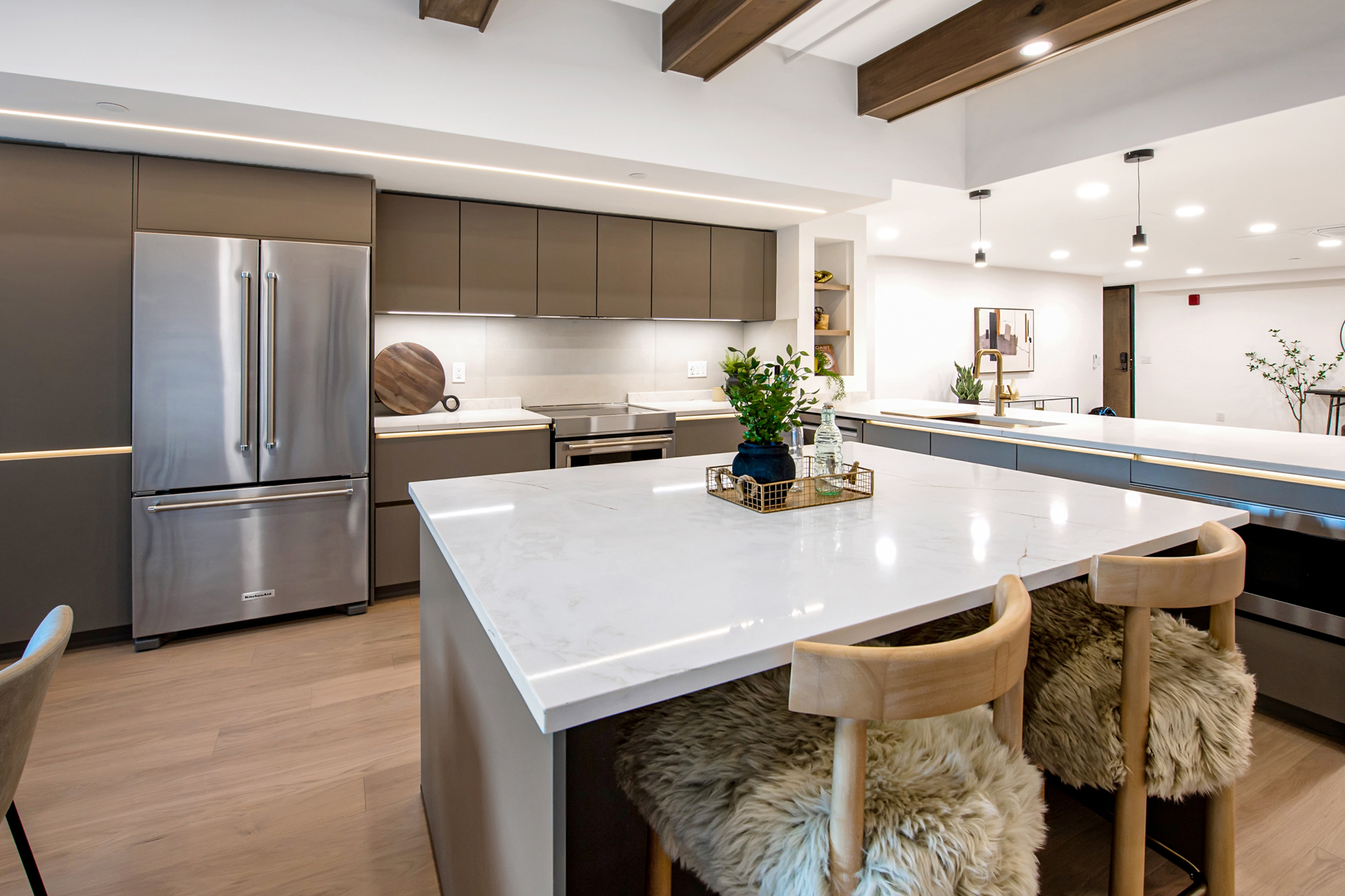 This modern, high-end kitchen features sleek, matte-finish cabinetry in a warm taupe tone paired with a large, white marble-veined quartz island. The space is accented by rustic wooden ceiling beams, warm under-cabinet lighting, and unique bar stools with faux-fur cushions, creating a sophisticated yet cozy atmosphere. The perspective is a wide-angle shot from the island, showcasing the seamless flow between the cooking area and the open-concept living space beyond.