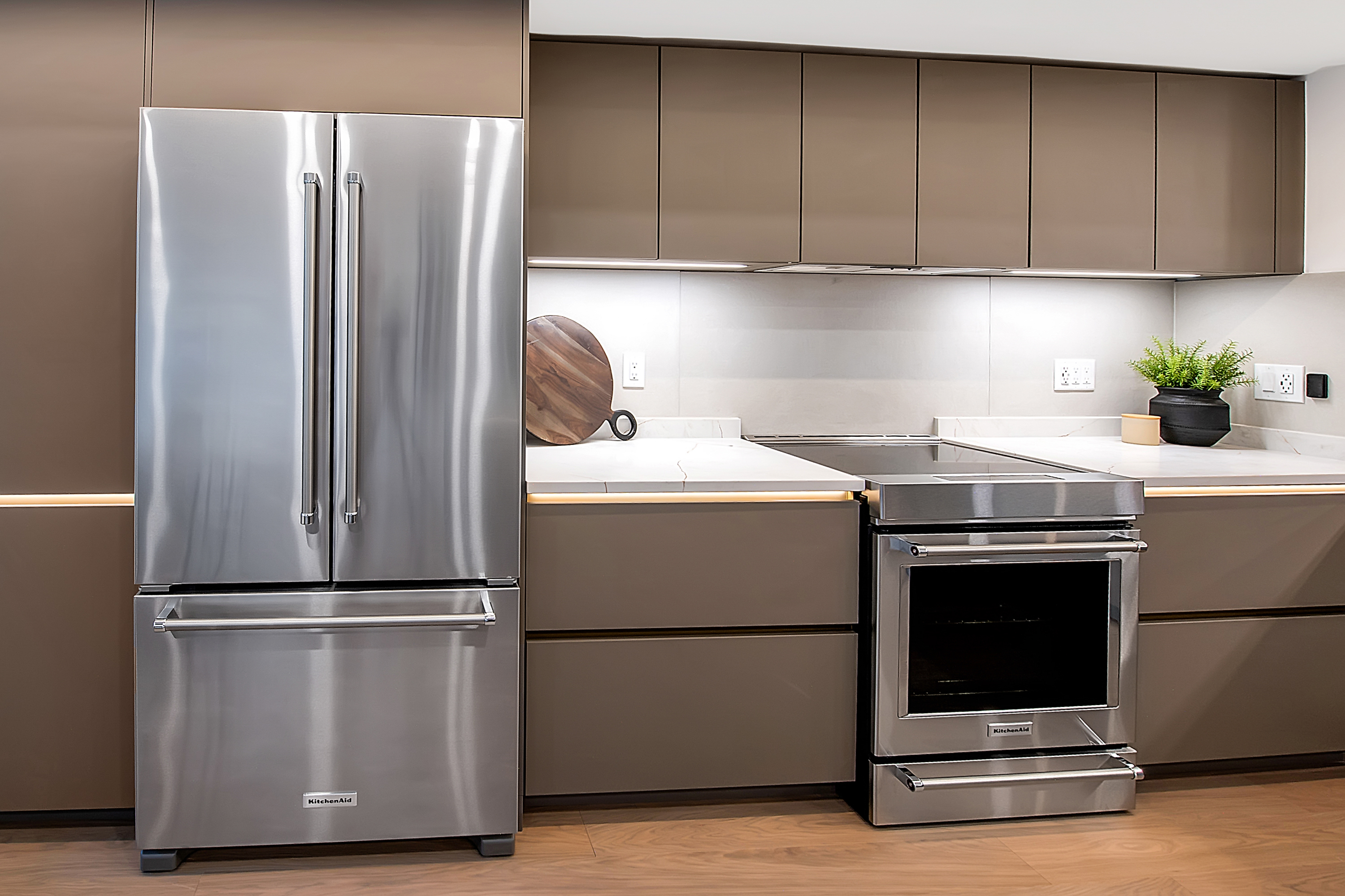 This sleek, modern kitchen features high-end stainless steel KitchenAid appliances, including a large French-door refrigerator and a slide-in range. The cabinetry is finished in a sophisticated matte taupe, complemented by white quartz countertops and integrated under-cabinet lighting that creates a warm, inviting atmosphere. The clean lines and minimalist aesthetic provide a contemporary and luxurious feel, perfect for a high-end real estate presentation.
