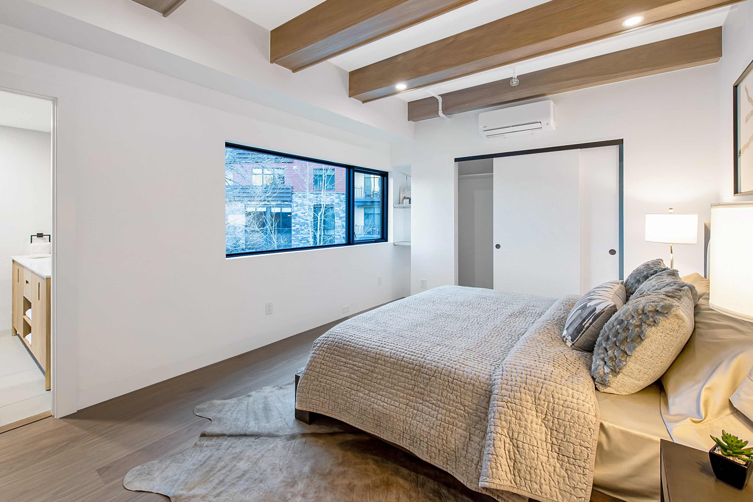This modern primary bedroom features a neutral color palette, warm wood ceiling beams, and a textured quilt on the bed. A large window provides natural light and a view of the exterior, while a sliding door leads to a closet and an adjacent bathroom is visible through the doorway. The room exudes a serene and contemporary atmosphere, enhanced by the soft lighting and minimalist decor.