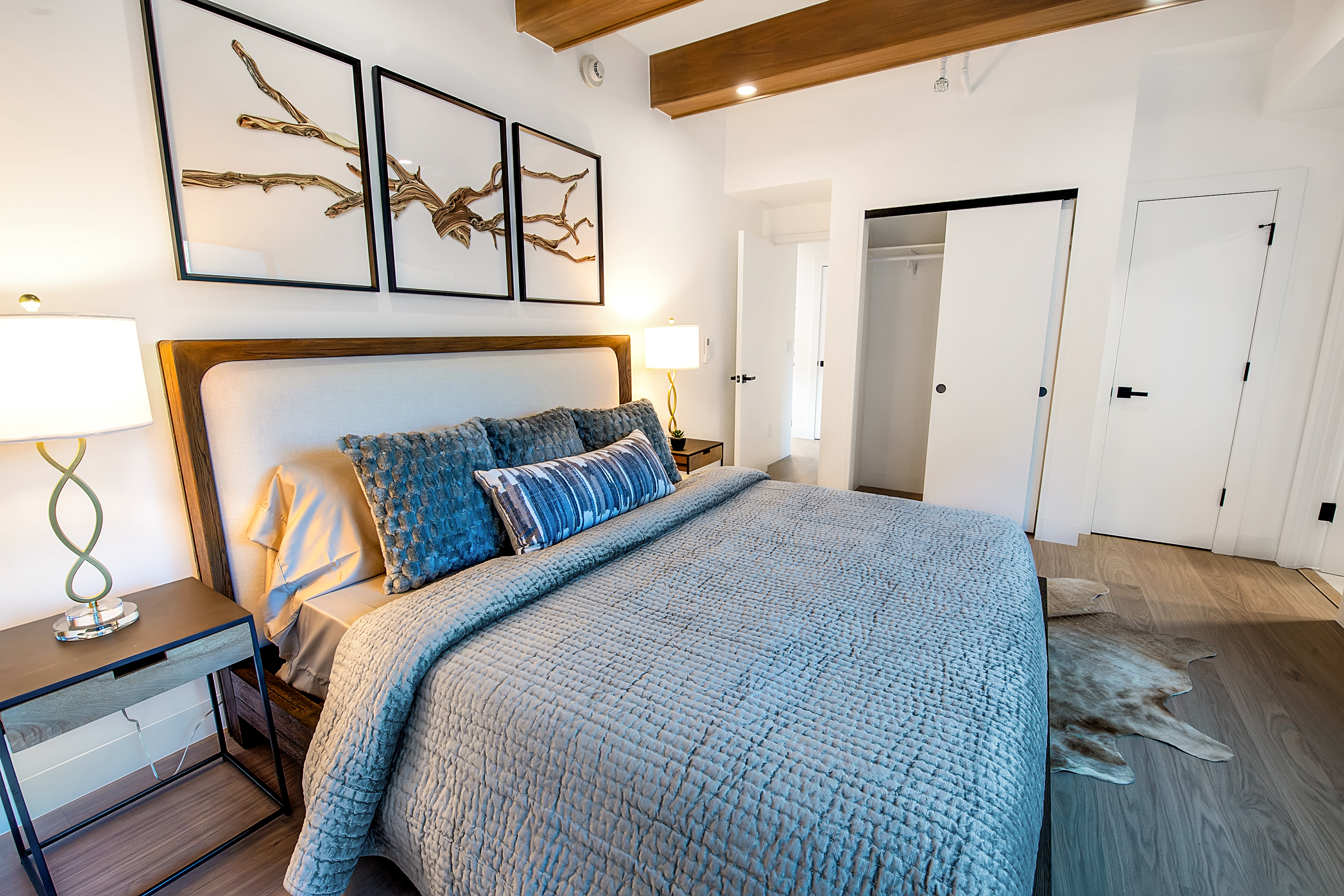 This modern primary bedroom features a plush, textured grey bedspread accented with blue decorative pillows and a matching lumbar pillow. Above the upholstered headboard, three framed pieces of driftwood art create a sophisticated focal point, while warm bedside lamps provide a cozy atmosphere. The room is finished with light wood-look flooring, a faux-hide rug, and exposed wooden ceiling beams, offering a clean and contemporary aesthetic.