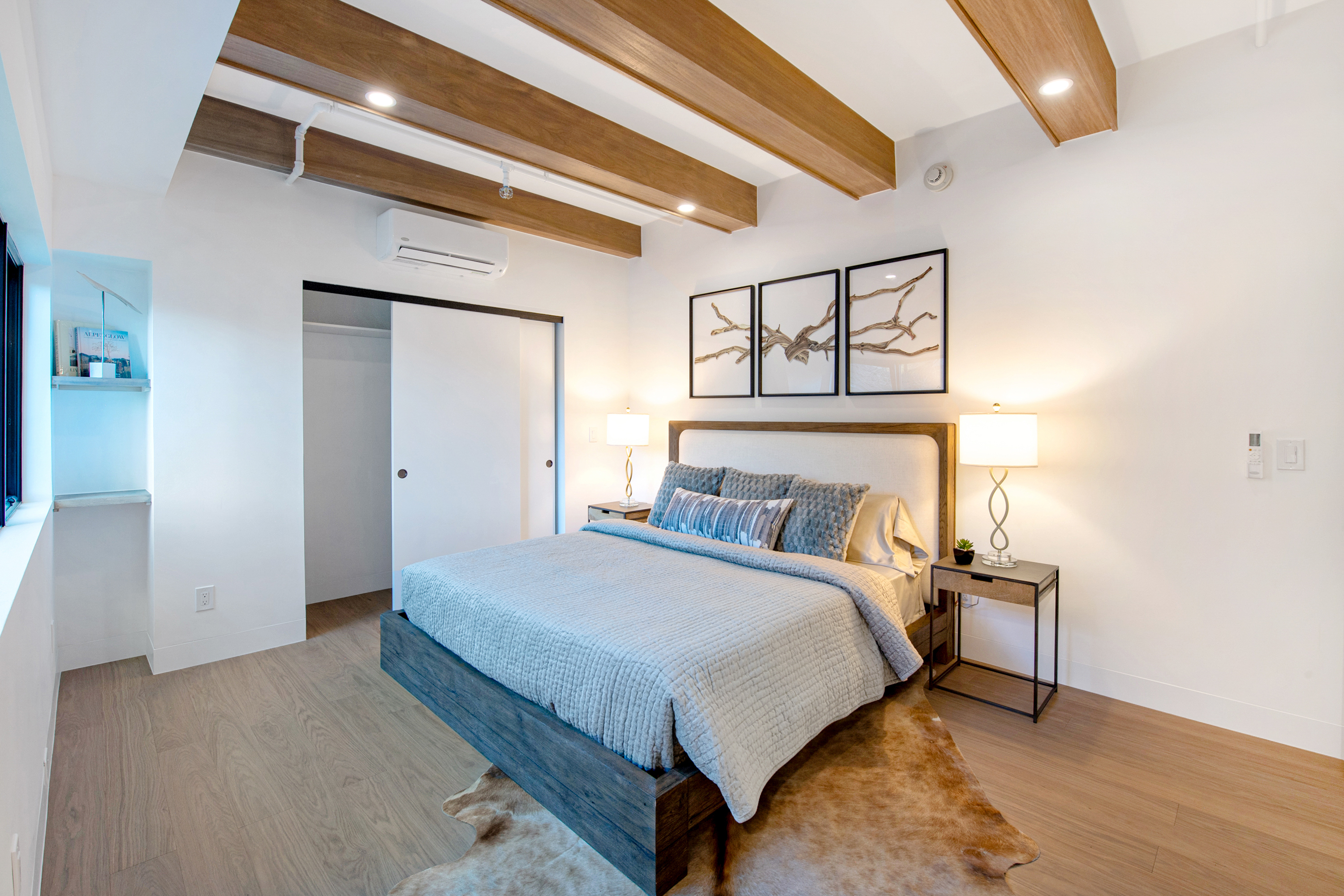 This modern primary bedroom features a platform bed with a textured grey quilt, framed by a wooden headboard and matching nightstands with elegant lamps. The room is accented by exposed wooden ceiling beams, a cowhide rug, and a triptych of branch-themed artwork above the bed. A sliding door leads to a closet, and a built-in shelving unit adds a functional, minimalist touch to the bright, airy space.