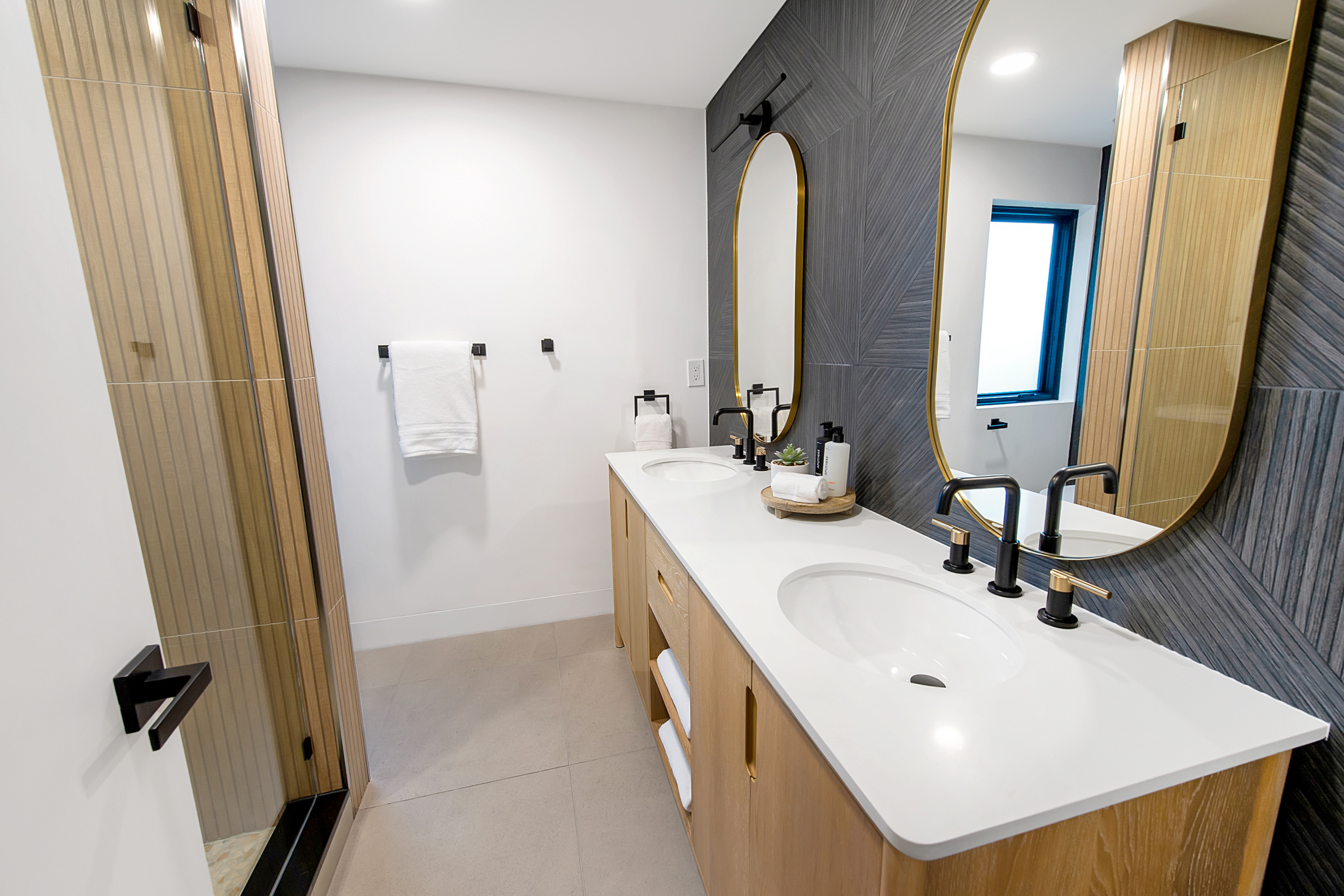 This modern primary bathroom features a sleek double vanity with light wood cabinetry, a white quartz countertop, and matte black fixtures. The space is accented by a striking dark gray geometric tiled wall and two elegant oval mirrors with gold frames, creating a sophisticated and contemporary aesthetic. The perspective captures the full length of the vanity, highlighting the clean lines and high-end finishes that define this luxurious bathroom.
