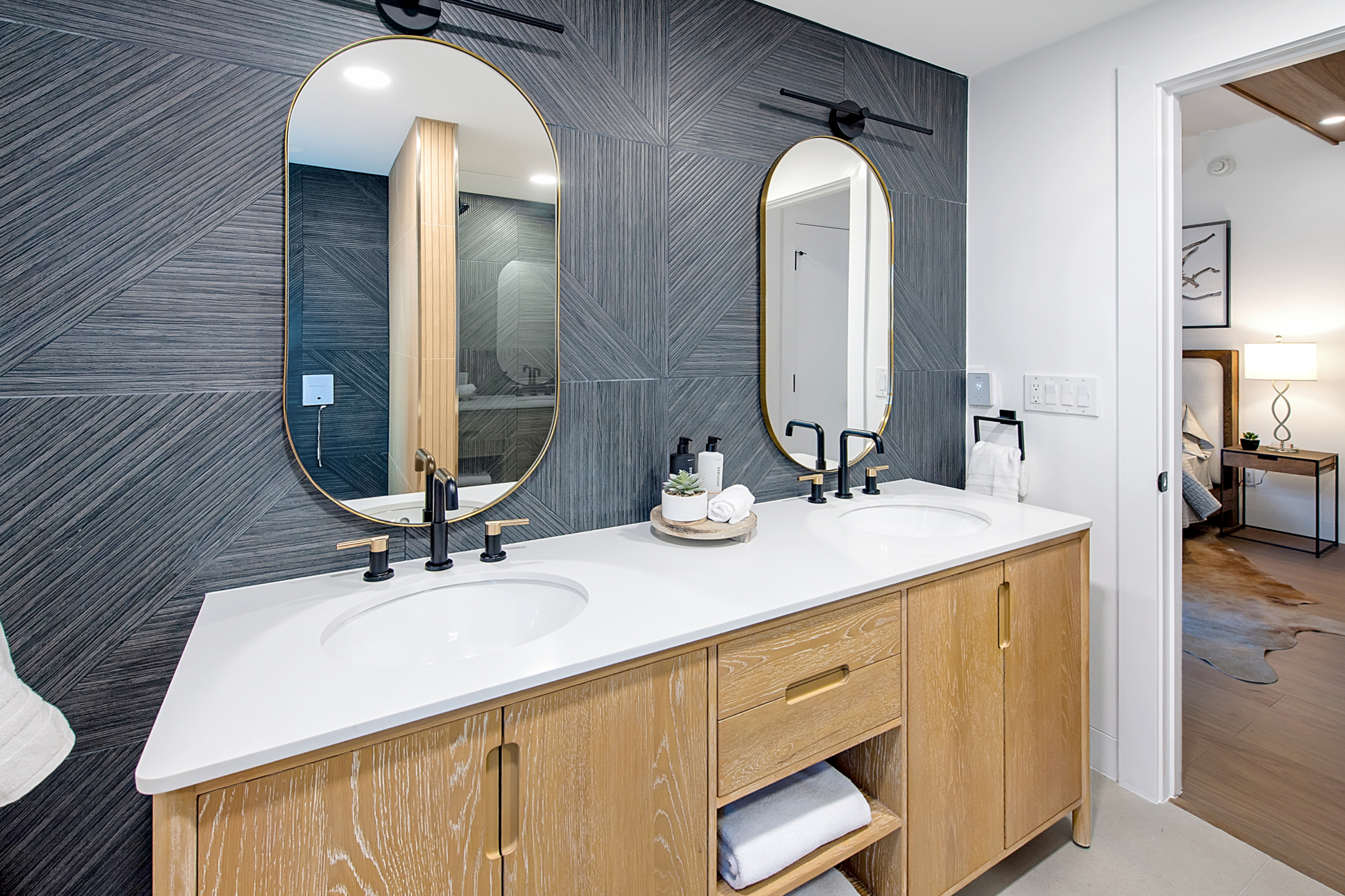 This modern primary bathroom features a dual-sink vanity with light oak cabinetry, white quartz countertops, and matte black fixtures. The wall is accented with dark, textured geometric tiles and two oval mirrors with gold frames, creating a sophisticated and contemporary aesthetic. A glimpse into the adjacent bedroom adds depth to the composition, highlighting the seamless flow of the home's design.