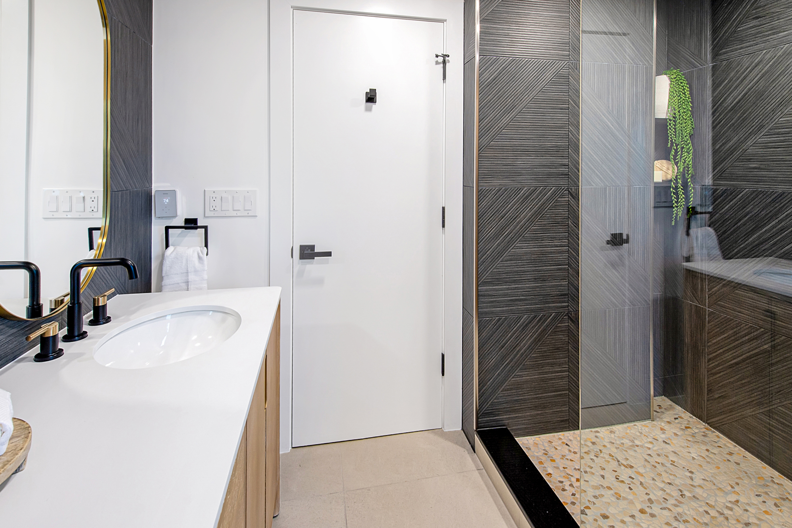 This modern bathroom features a sleek vanity with a white countertop, a black faucet, and a gold-rimmed mirror. To the right, a walk-in shower is enclosed by glass and showcases striking, textured dark gray geometric tiles and a pebble-stone floor. The space is bright and contemporary, blending high-contrast materials with clean lines for a sophisticated, spa-like aesthetic.