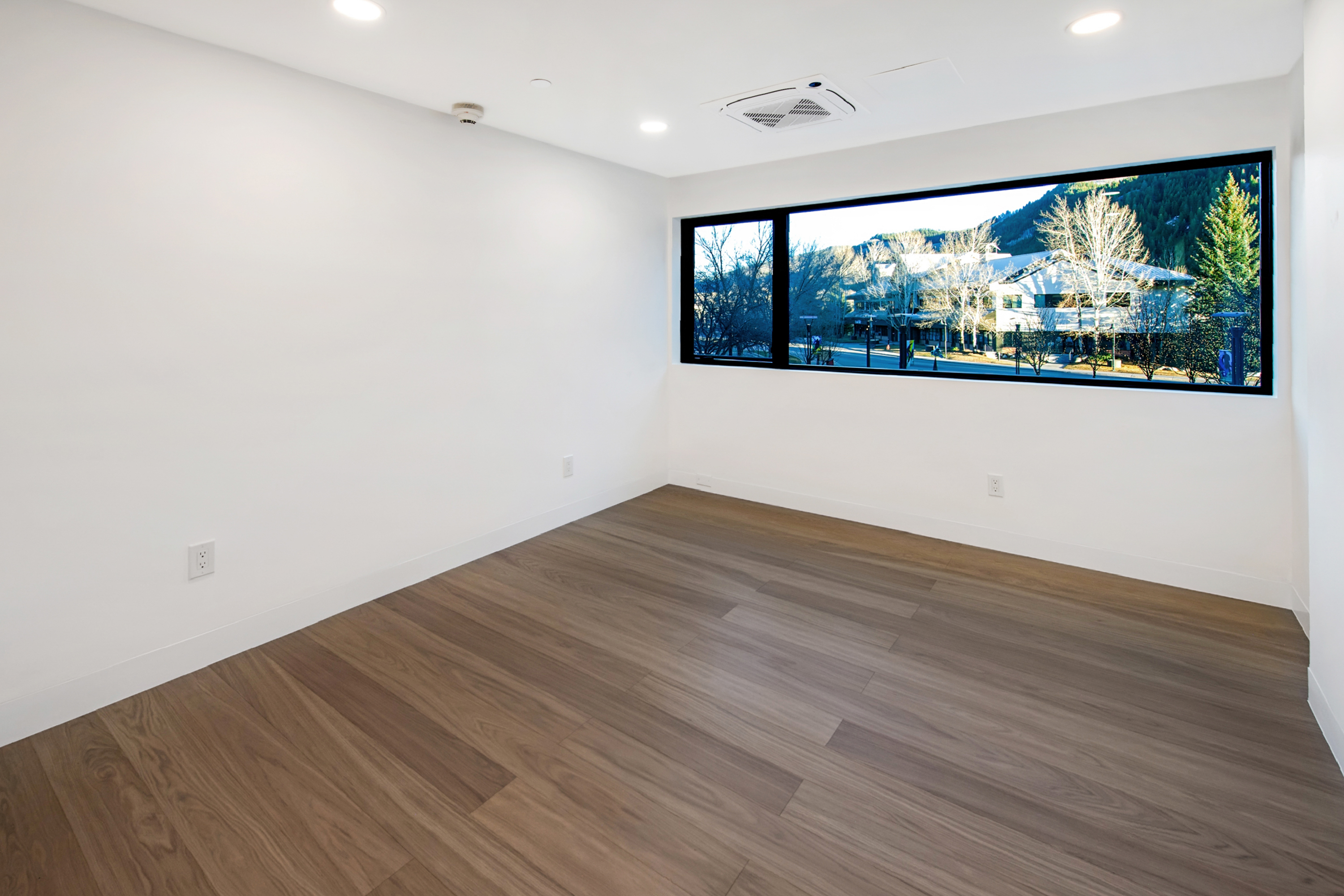 This minimalist, unfurnished room features clean white walls, light-toned wood-look flooring, and a wide, horizontal window that frames a scenic view of trees and buildings. The space is illuminated by recessed ceiling lights and a central HVAC unit, creating a bright, versatile environment suitable for a home office or study. The perspective is from the corner of the room, emphasizing the open floor plan and the natural light pouring in from the exterior.