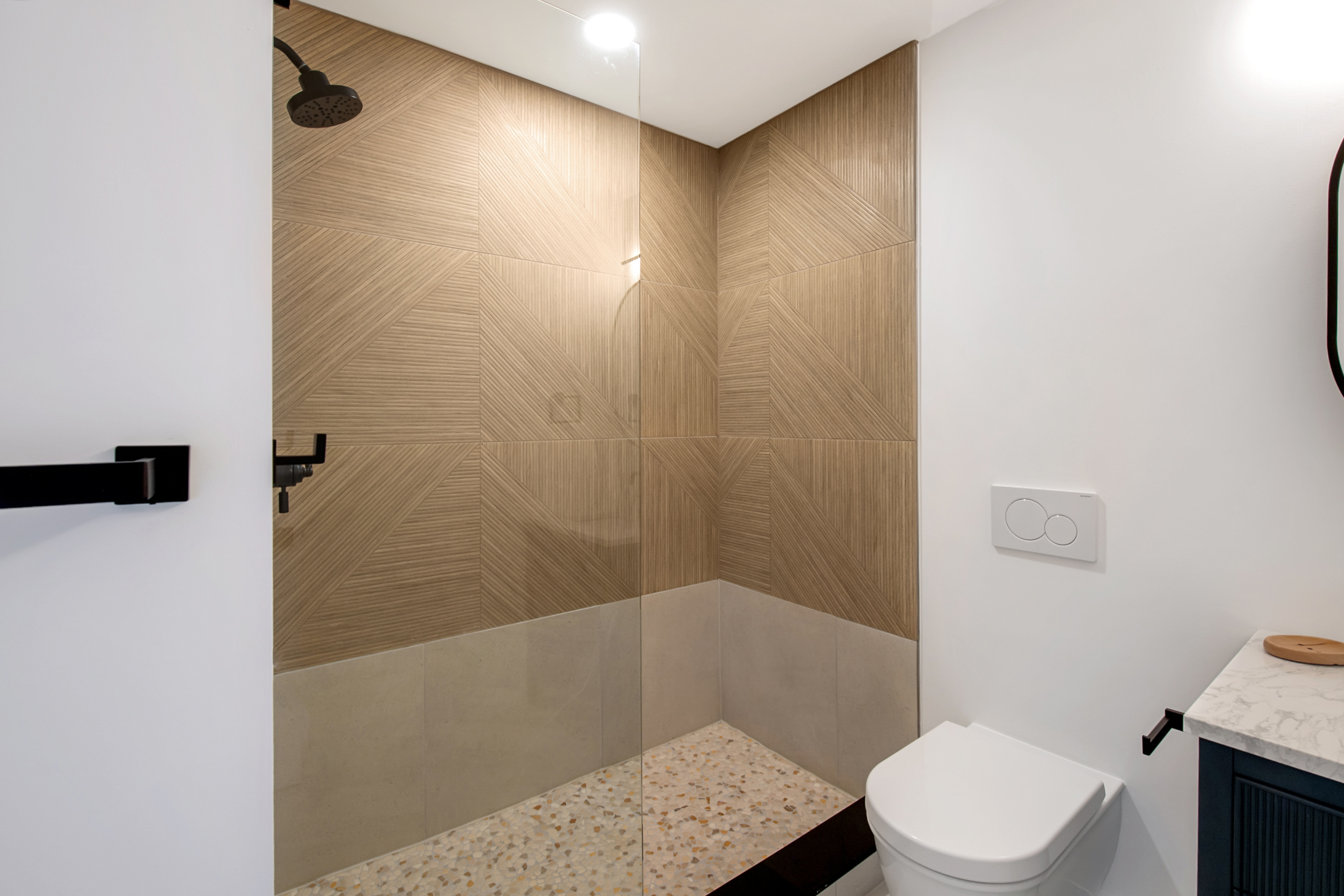 This modern bathroom features a walk-in shower with a striking geometric-patterned tile accent wall and a light-colored terrazzo floor. A sleek glass partition separates the shower from the rest of the room, which includes a wall-mounted toilet and a vanity with a marble countertop. The minimalist design, combined with high-contrast black fixtures, creates a sophisticated and contemporary aesthetic.