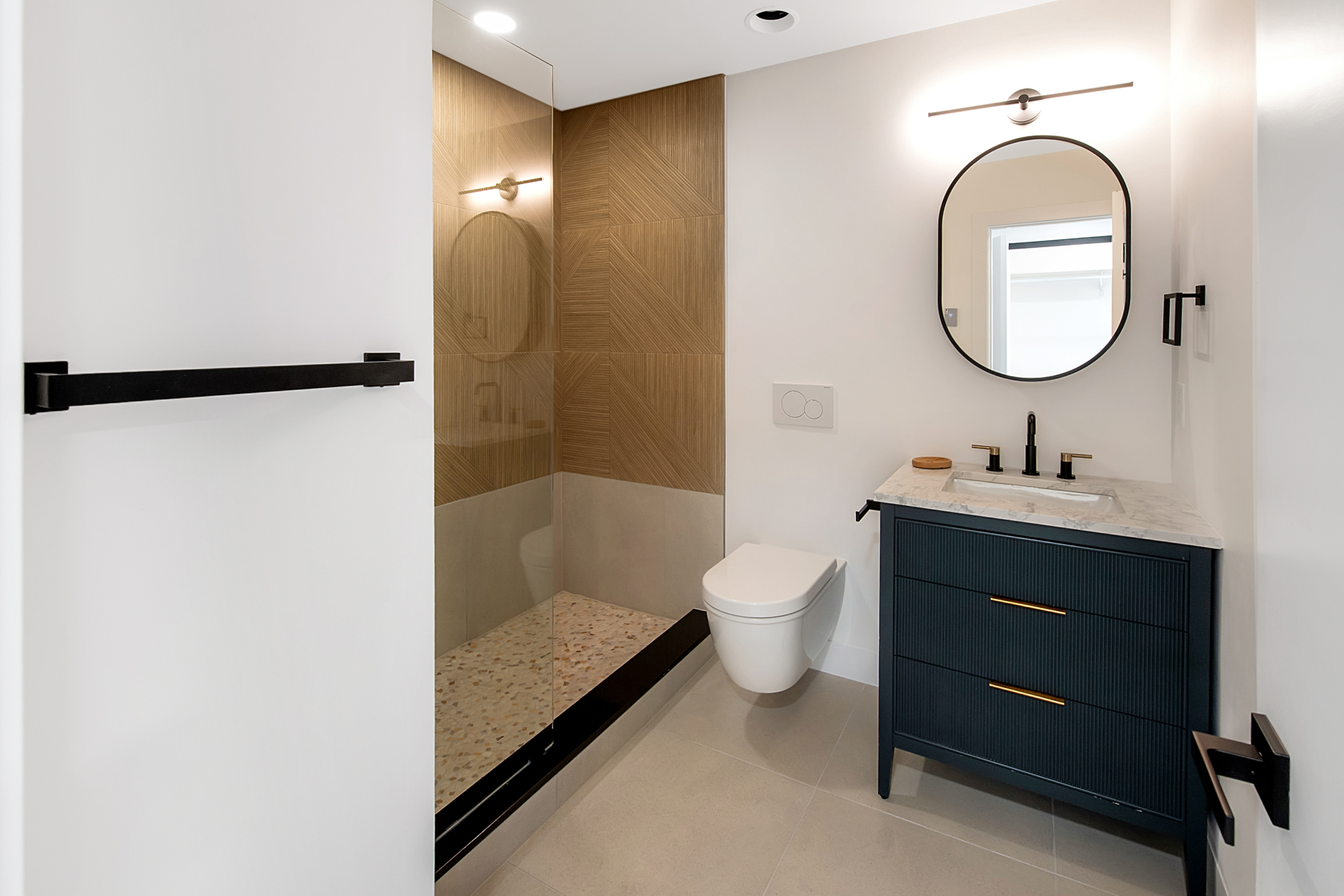 This modern bathroom features a sleek, dark blue vanity with gold hardware, topped with a marble countertop and a minimalist oval mirror. To the left, a walk-in shower with a glass partition showcases textured wood-look wall tiles and a speckled stone floor, complemented by a wall-mounted toilet. The overall aesthetic is contemporary and sophisticated, utilizing a high-contrast color palette and clean lines to create a luxurious feel.