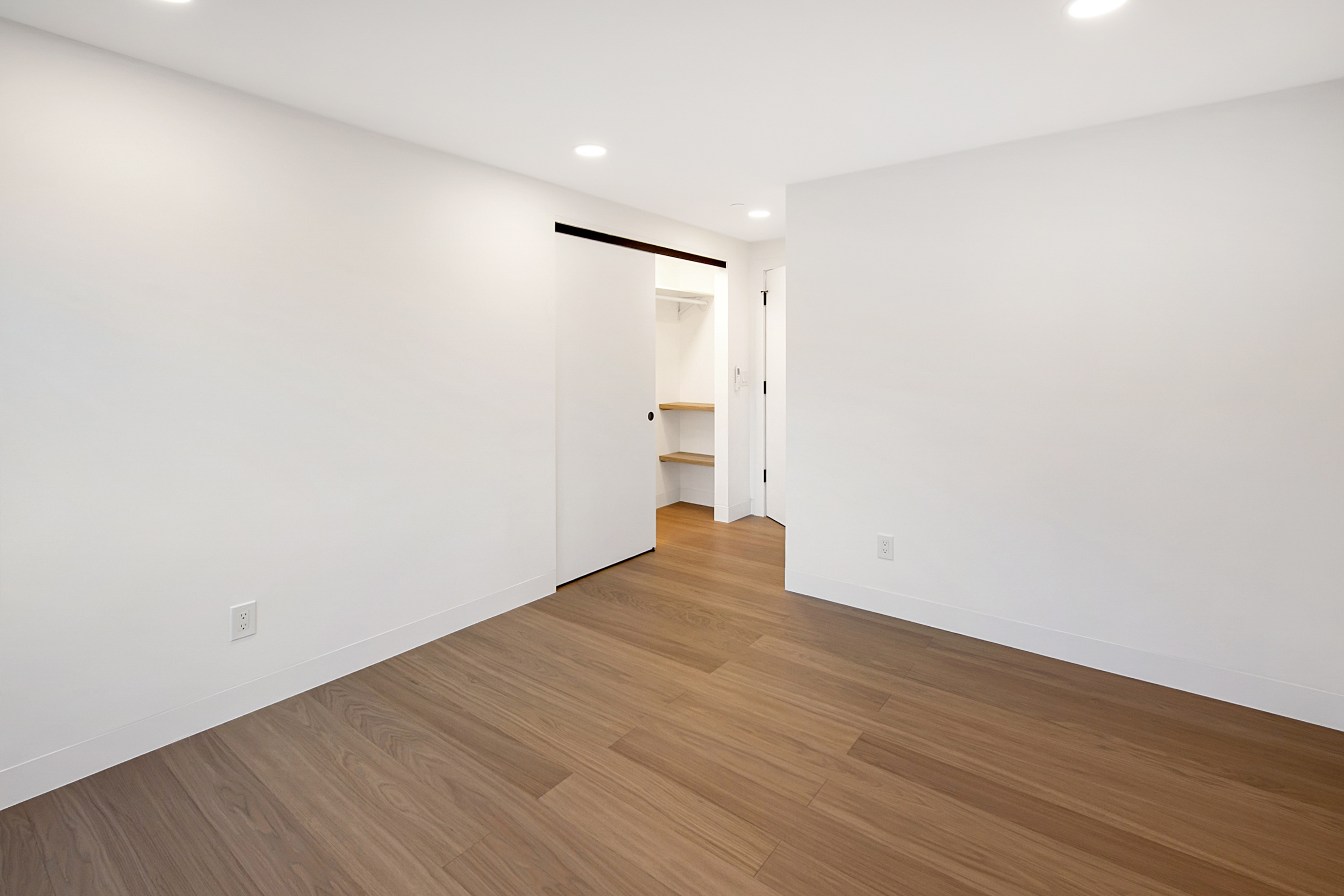 This minimalist, unfurnished room features clean white walls and warm-toned wood-look flooring, creating a bright and versatile space. A sliding door reveals a walk-in closet with built-in shelving, while recessed lighting provides even illumination throughout. The perspective is centered, highlighting the open layout and the transition into the closet area.