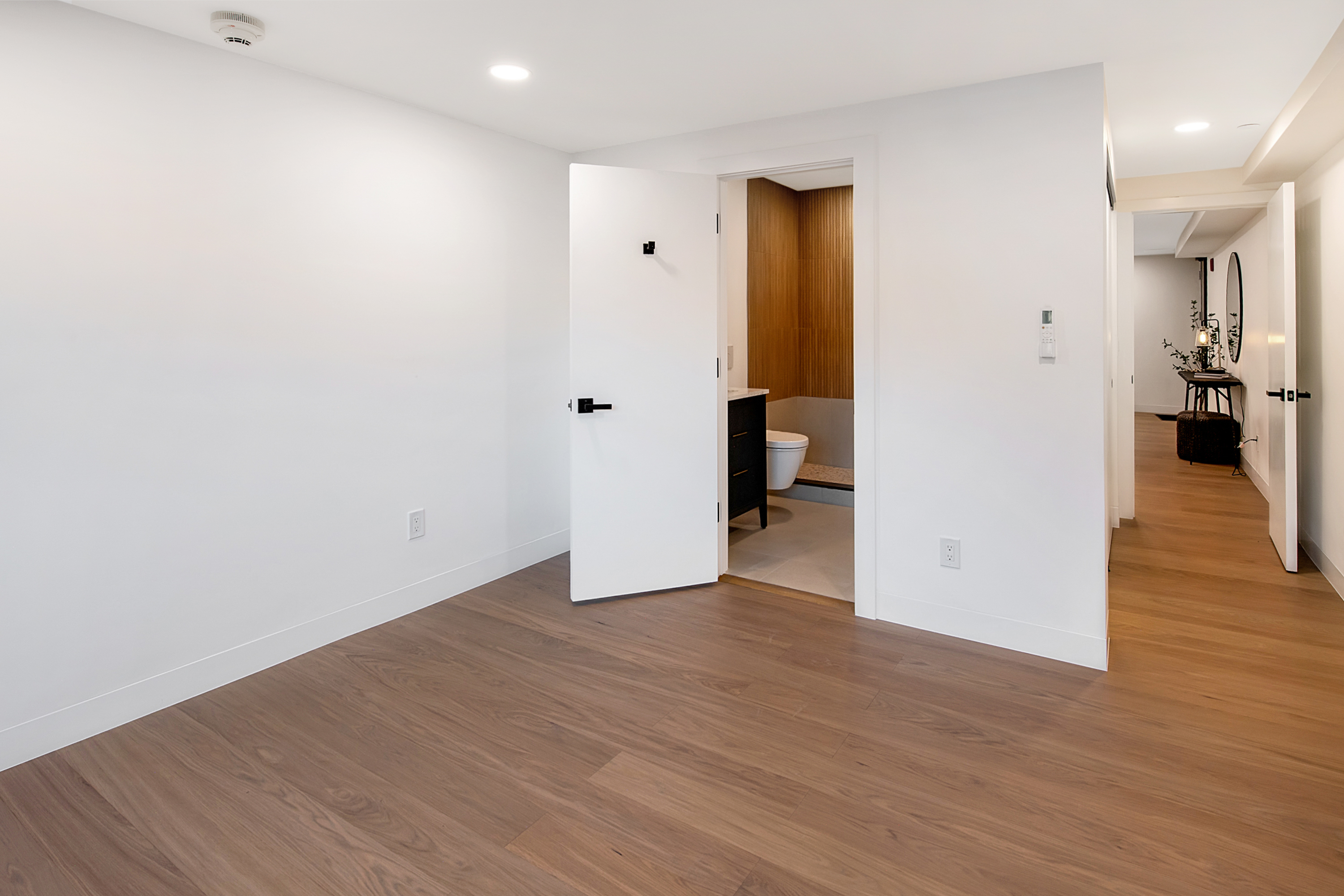 This interior view captures a clean, minimalist hallway space featuring light wood-look flooring and crisp white walls. The perspective looks into a small bathroom with wood-slat accent walls and continues down a corridor toward a secondary room with a console table and mirror. The overall aesthetic is modern, bright, and neutral, emphasizing a seamless flow between living areas.