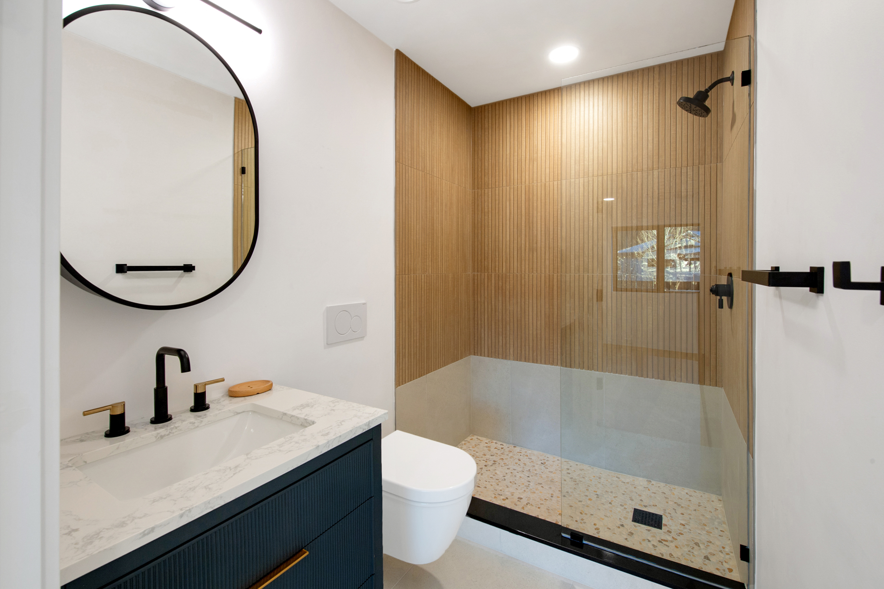 This modern bathroom features a sleek vanity with a marble countertop, a dark blue fluted cabinet, and a striking round black-framed mirror. The walk-in shower is a standout, showcasing vertical wood-look tile walls, a terrazzo-style floor, and a glass enclosure with matte black fixtures. The space is bright and contemporary, blending natural textures with bold, clean lines for a sophisticated aesthetic.
