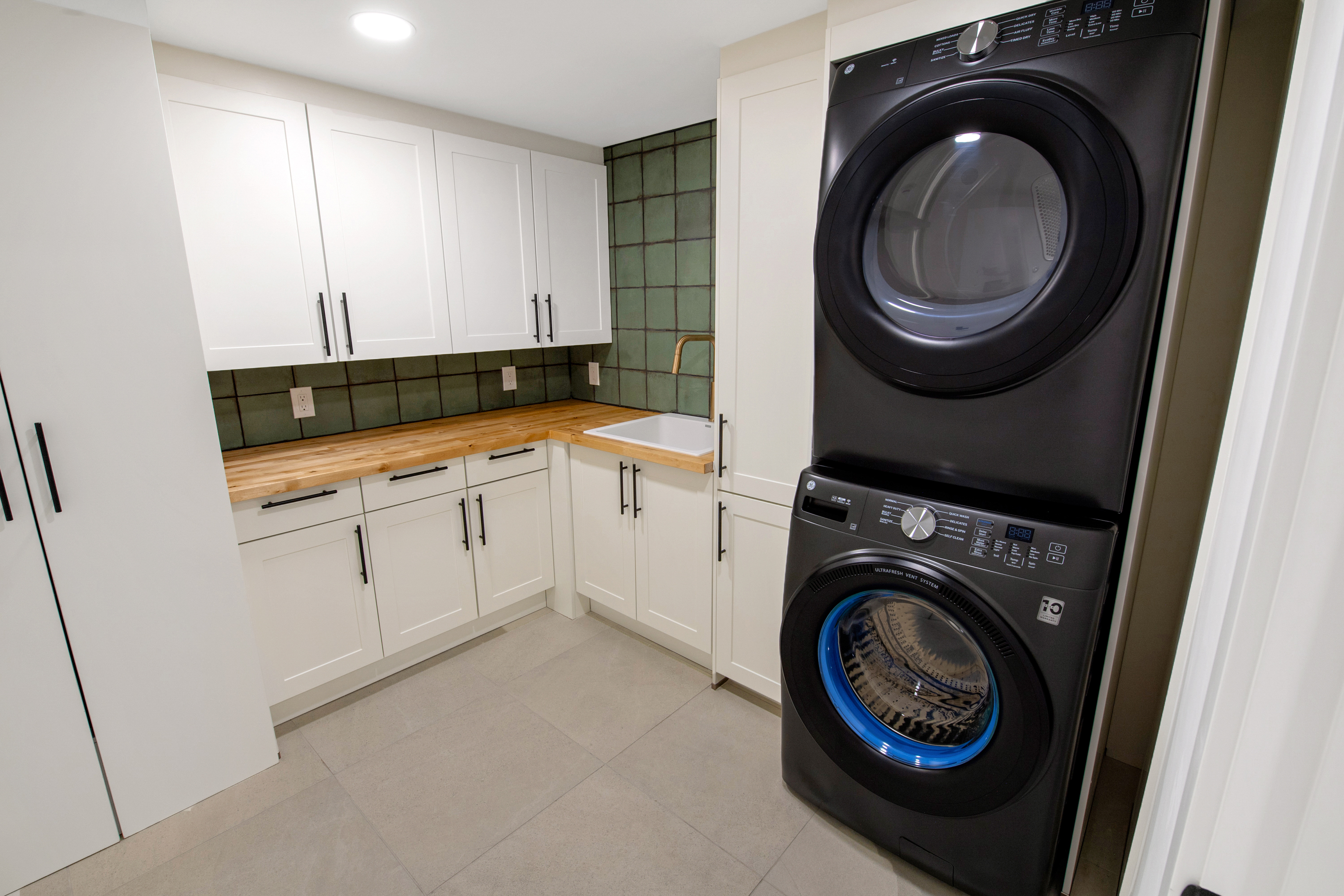 This modern laundry room features a sleek, stacked black washer and dryer set contrasted against crisp white cabinetry and a warm wood countertop. A deep white utility sink with a gold faucet is set against a striking green tiled backsplash, creating a clean and functional workspace. The room is well-lit with recessed lighting and finished with neutral floor tiles, offering a contemporary and organized aesthetic.