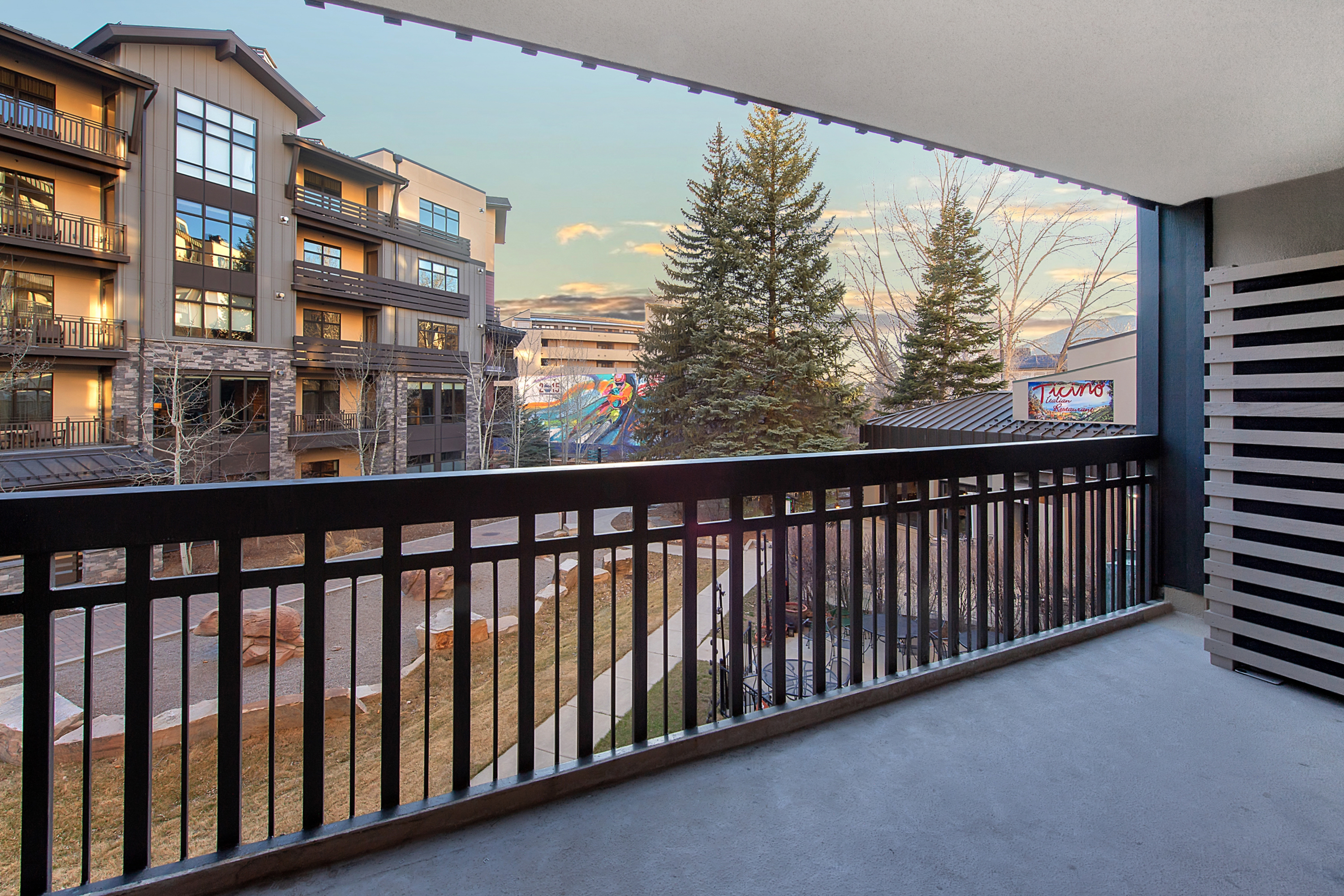 This balcony offers a clear, elevated view of a modern multi-story residential building and a nearby restaurant with a prominent sign. The space features a sleek black metal railing and a concrete floor, providing a clean and contemporary outdoor area. The perspective captures the surrounding landscape, including trees and a mural, creating an inviting atmosphere for relaxation.