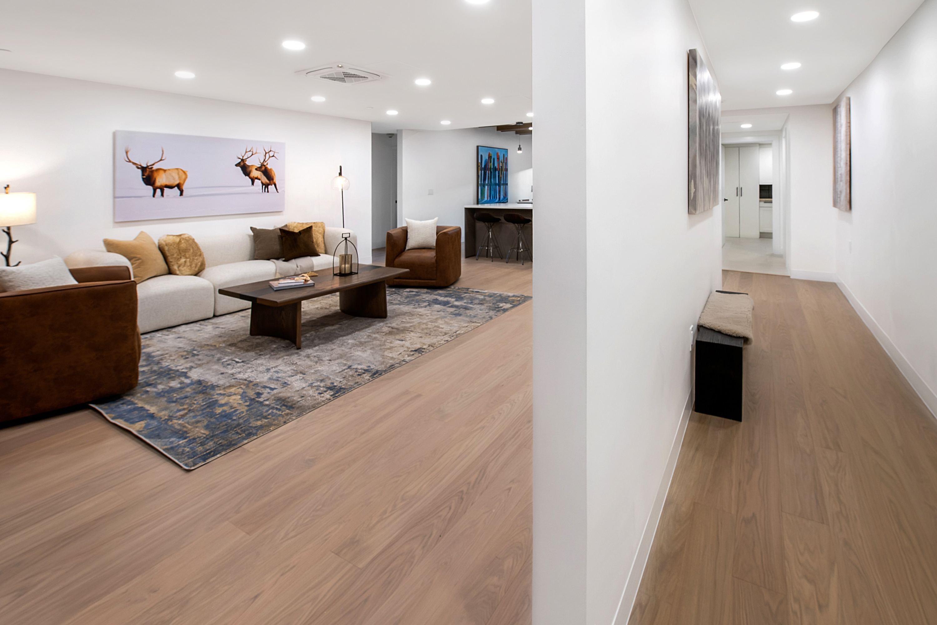 This open-concept living area features a sophisticated blend of modern design and warm, earthy tones, anchored by a large, textured area rug and a sleek wooden coffee table. The space is furnished with a plush white sofa and rich brown leather armchairs, complemented by striking elk-themed wall art and recessed lighting. A smooth transition leads from the cozy lounge area into a bright, minimalist hallway, creating a seamless and inviting flow throughout the home.
