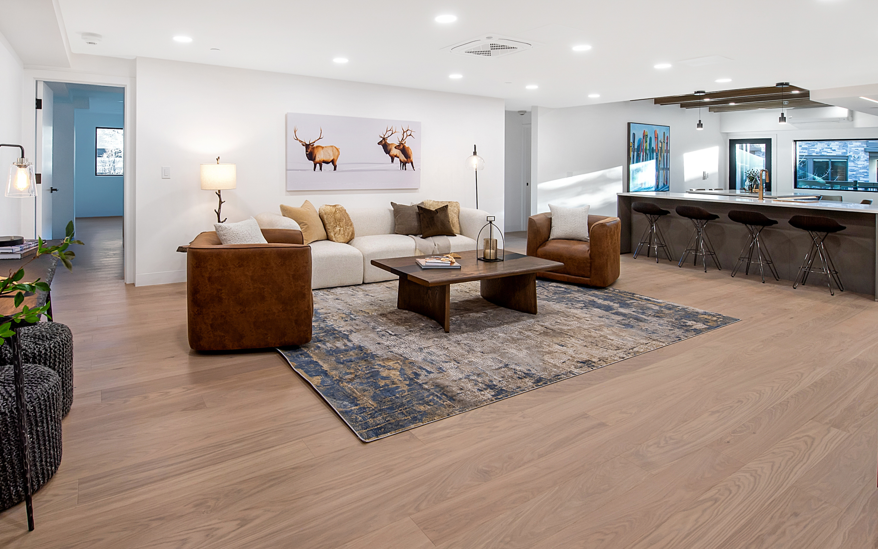 This spacious, open-concept living area features a contemporary design with light hardwood flooring and a neutral color palette. The room is anchored by a large, textured area rug, a plush cream sofa, and two leather armchairs, all centered around a substantial wooden coffee table. A large elk-themed wall art piece serves as a focal point, while the adjacent kitchen area with a breakfast bar creates a seamless flow for entertaining.