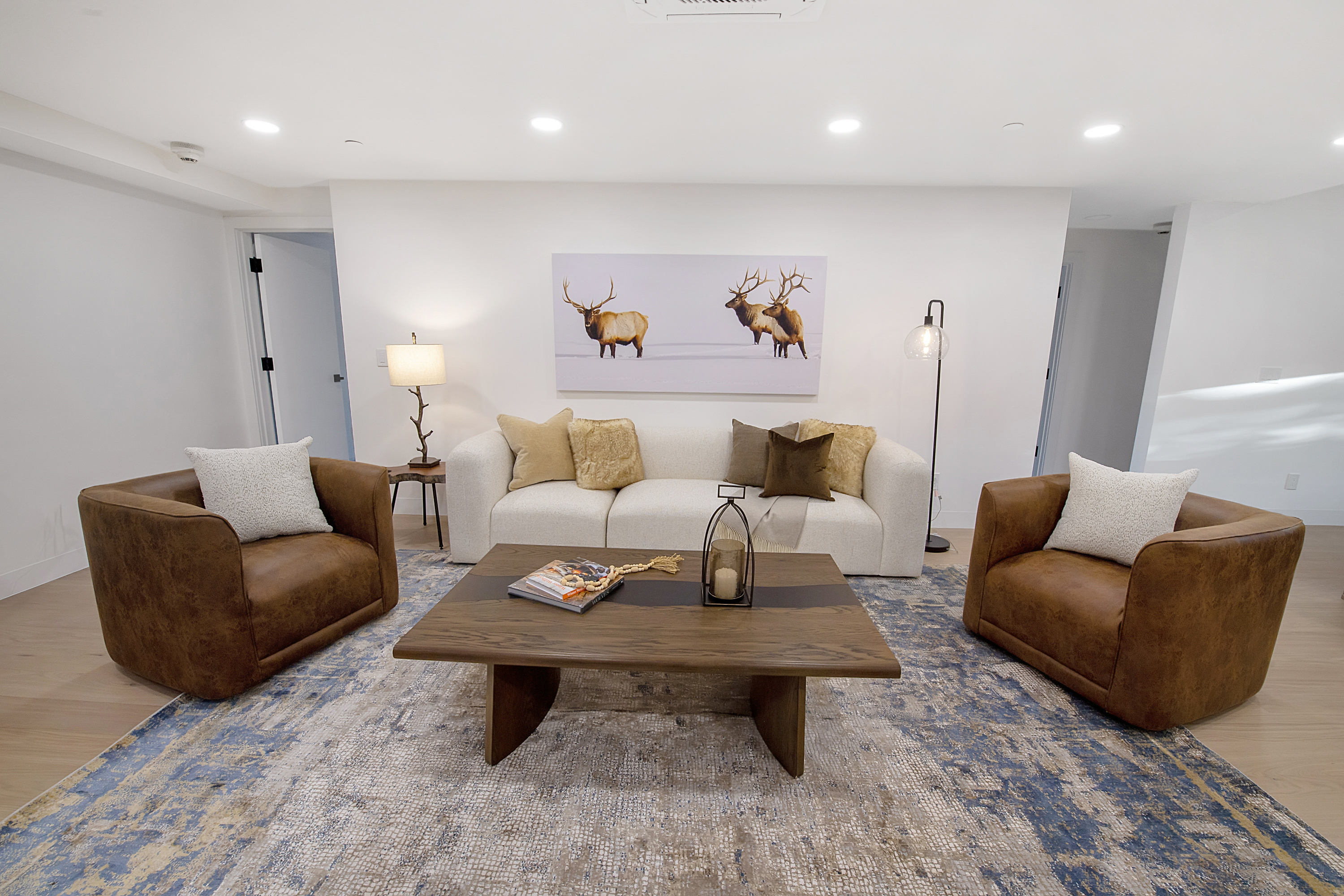 This inviting living room features a symmetrical layout with a plush white sofa centered beneath a large elk-themed wall art piece, flanked by two rich brown leather armchairs. A substantial wooden coffee table with a dark inlay serves as the focal point, resting on a textured blue and cream area rug that anchors the space. The room is illuminated by recessed ceiling lights, a sleek floor lamp, and a decorative table lamp, creating a warm and sophisticated atmosphere.