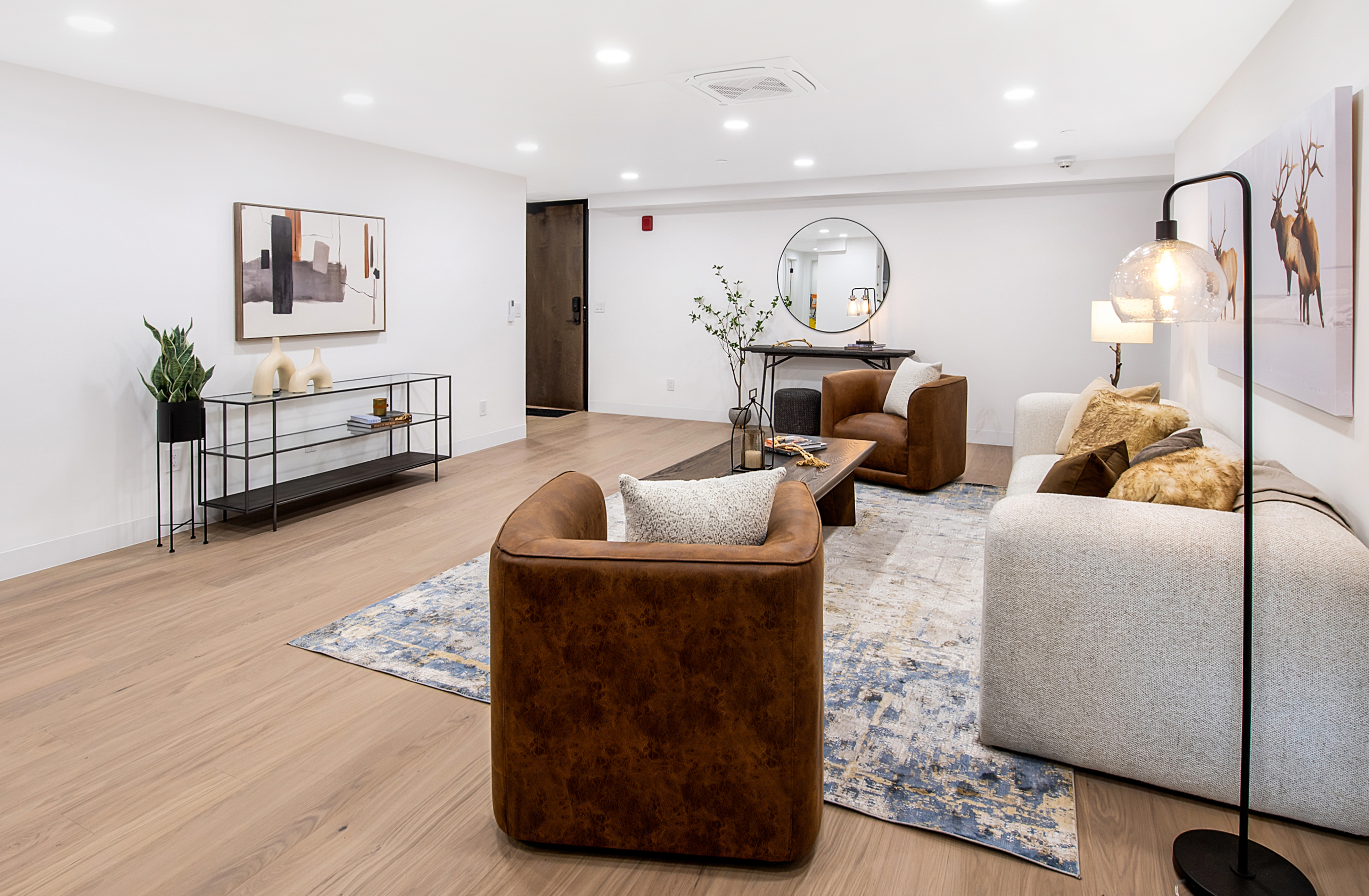This spacious and modern living room features a warm, inviting aesthetic with light wood flooring and a neutral color palette. The room is anchored by a large, textured area rug, a plush light-colored sofa, and two rich brown leather armchairs, creating a comfortable conversation area. A sleek glass-and-metal console table, a circular wall mirror, and contemporary artwork add sophisticated touches to this well-lit, open-concept space.