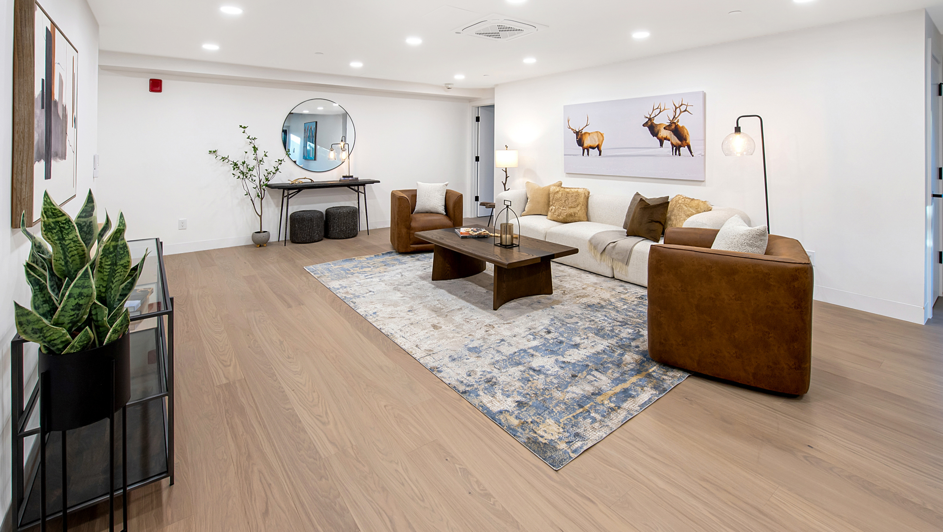 This spacious and modern living room features a neutral color palette with warm leather accents, centered around a large, textured area rug and a rustic wooden coffee table. The room is anchored by a long, cream-colored sofa and two leather armchairs, complemented by a large elk-themed wall art piece and a circular mirror above a console table. The perspective is a wide-angle shot that captures the open, inviting layout, highlighting the clean lines and contemporary aesthetic of the space.