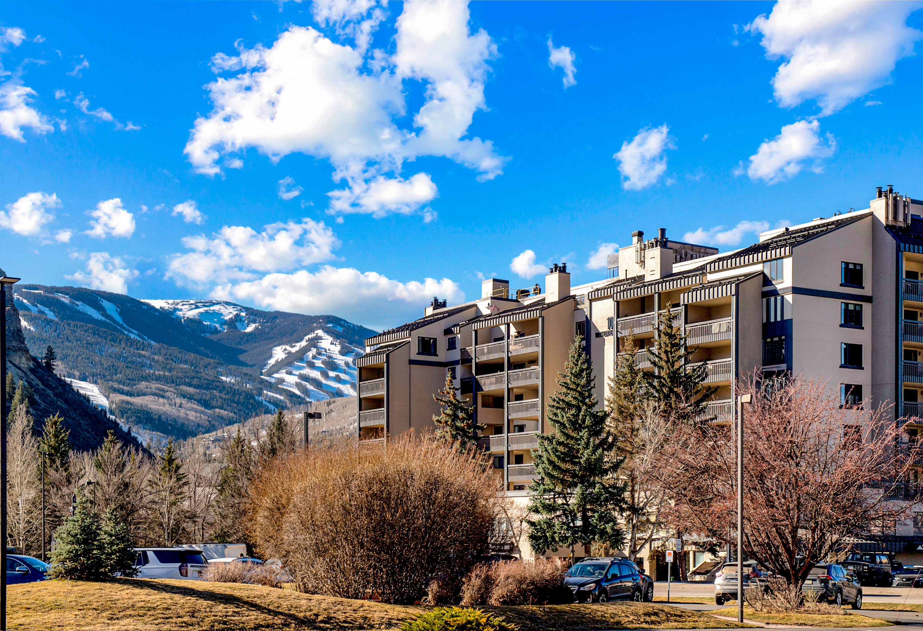 This exterior view showcases a multi-story residential complex with a modern, mountain-inspired architectural style, featuring balconies and a neutral color palette. The building is set against a dramatic backdrop of snow-capped mountains under a bright blue sky with scattered clouds. In the foreground, a landscaped area with trees and a parking lot provides a sense of scale and accessibility to the property.