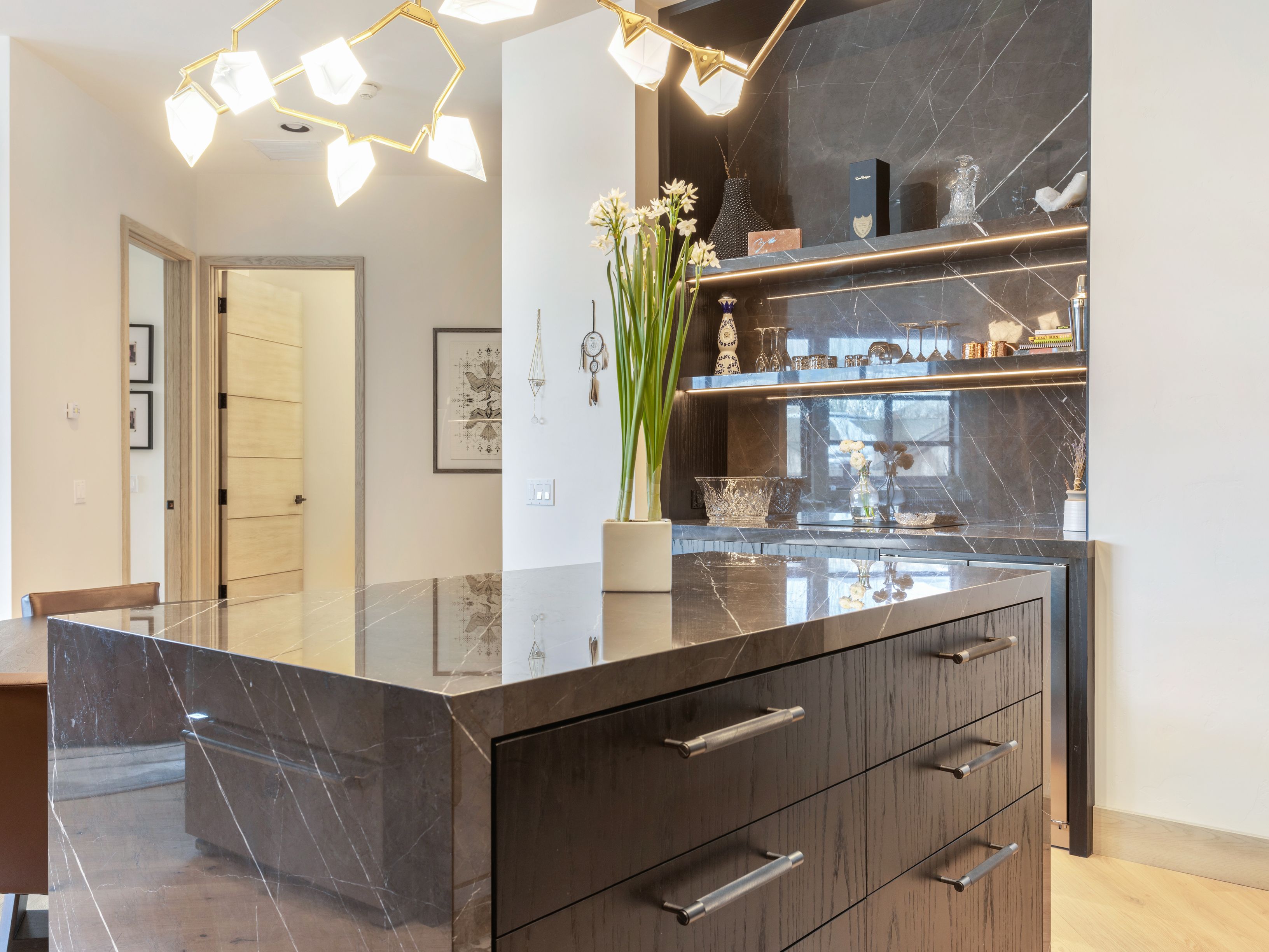 This interior shot showcases a modern kitchen island with dark wood cabinetry and a striking marble countertop. The island features sleek, silver hardware and is complemented by a built-in bar area with illuminated shelving displaying glassware and decorative items. A contemporary chandelier hangs above, adding a touch of elegance to the space.