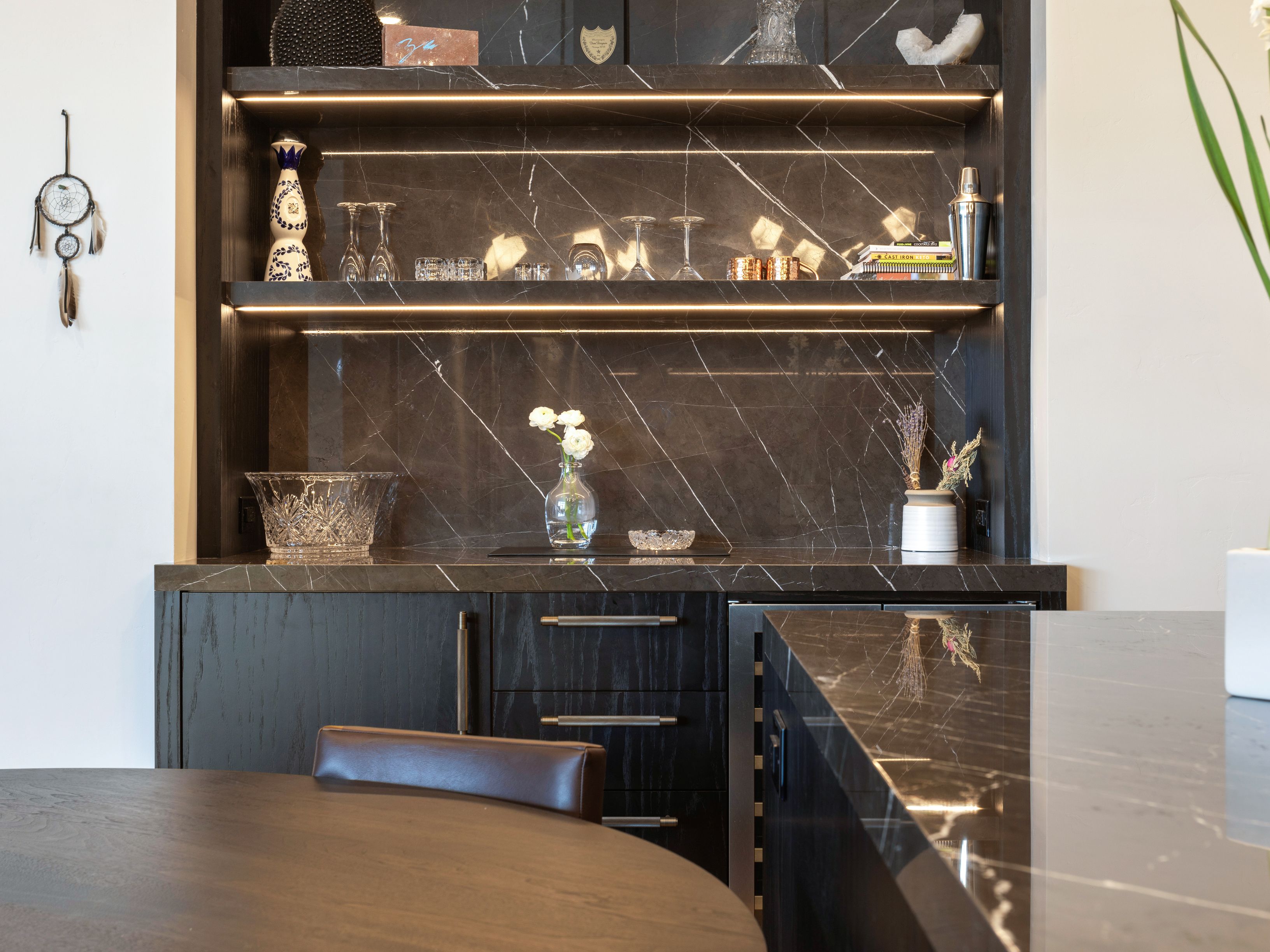 This image showcases a sophisticated dining area with a built-in bar featuring dark marble-like surfaces and illuminated shelving. The bar displays glassware and decorative items, adding a touch of elegance. A round wooden table and a leather chair are partially visible, suggesting a cozy and stylish dining space.