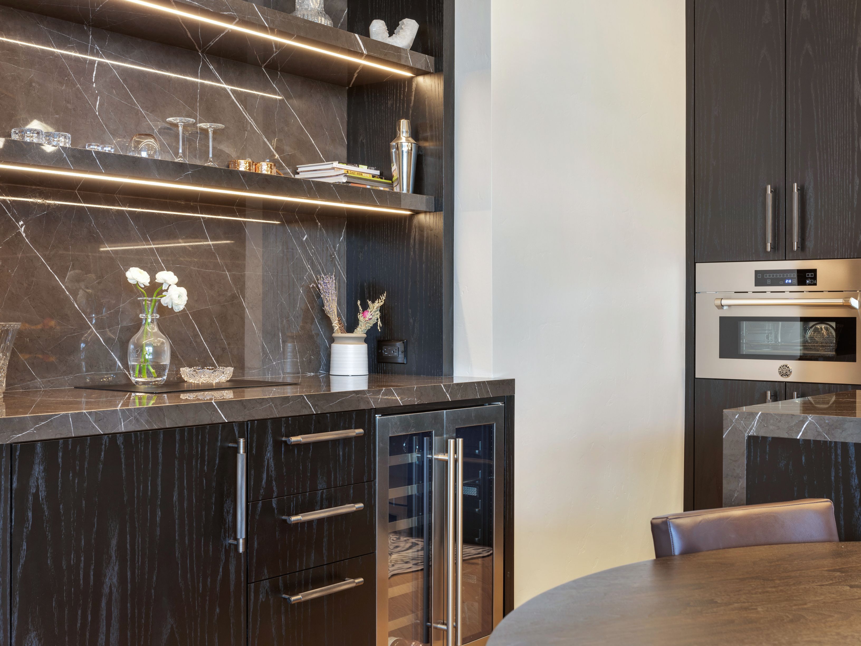This interior shot showcases a sophisticated kitchen area with dark wood cabinetry and marble countertops. The built-in shelving features integrated lighting, highlighting decorative items and glassware. A stainless steel oven and a wine cooler are seamlessly integrated into the design, adding to the luxurious feel of the space.