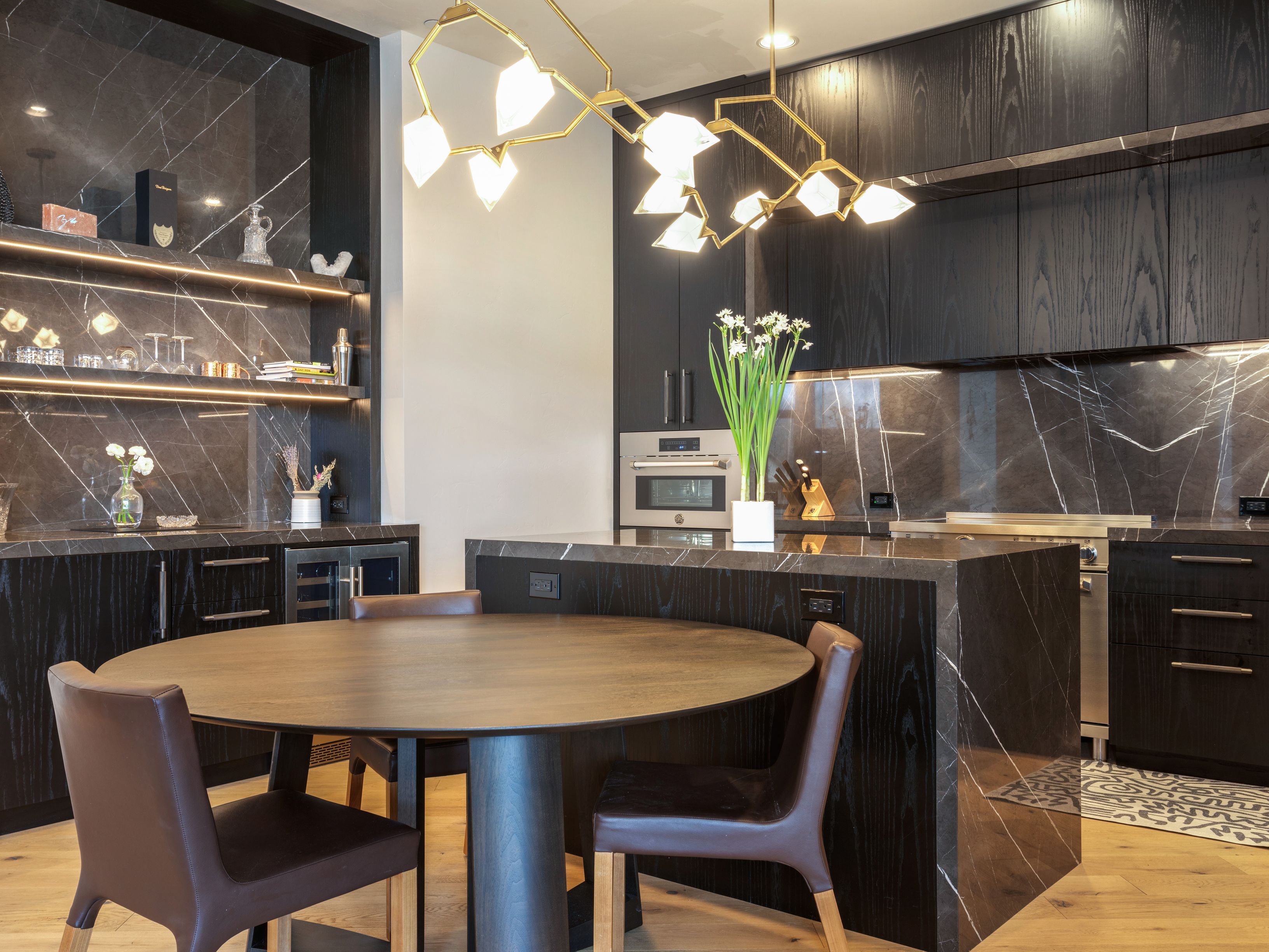 This is a modern kitchen featuring dark wood cabinetry and marble countertops with striking white veining. A unique, multi-bulb chandelier hangs above a round wooden dining table with brown leather chairs. The kitchen also includes a built-in wine cooler and open shelving with decorative items, creating a sophisticated and inviting space.