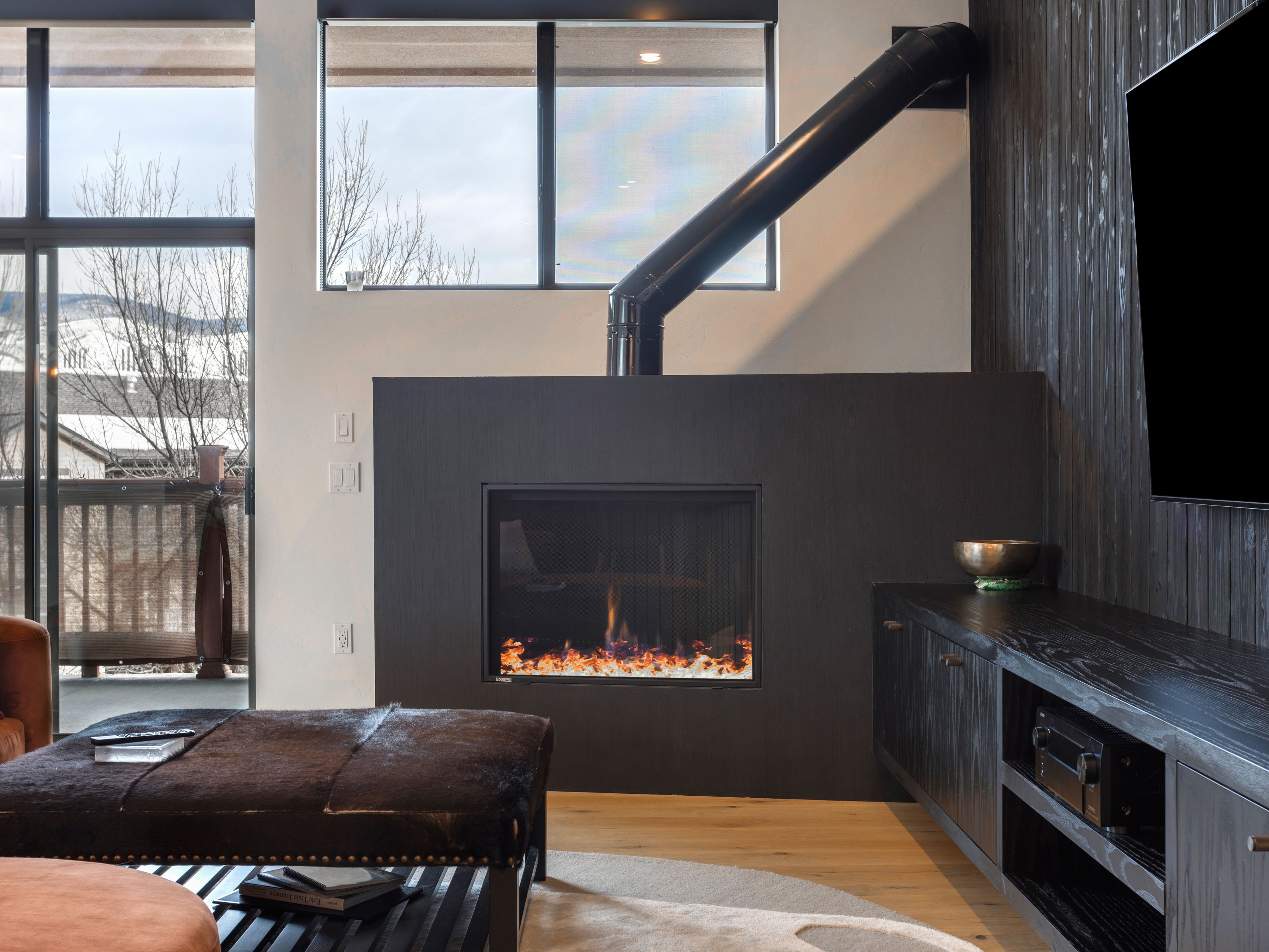 This is an interior shot of a modern living room featuring a dark wood fireplace with a black pipe extending upwards, a dark wood entertainment center, and a large ottoman. The room has large windows providing natural light and a view of the outdoors. The overall aesthetic is contemporary and sophisticated.