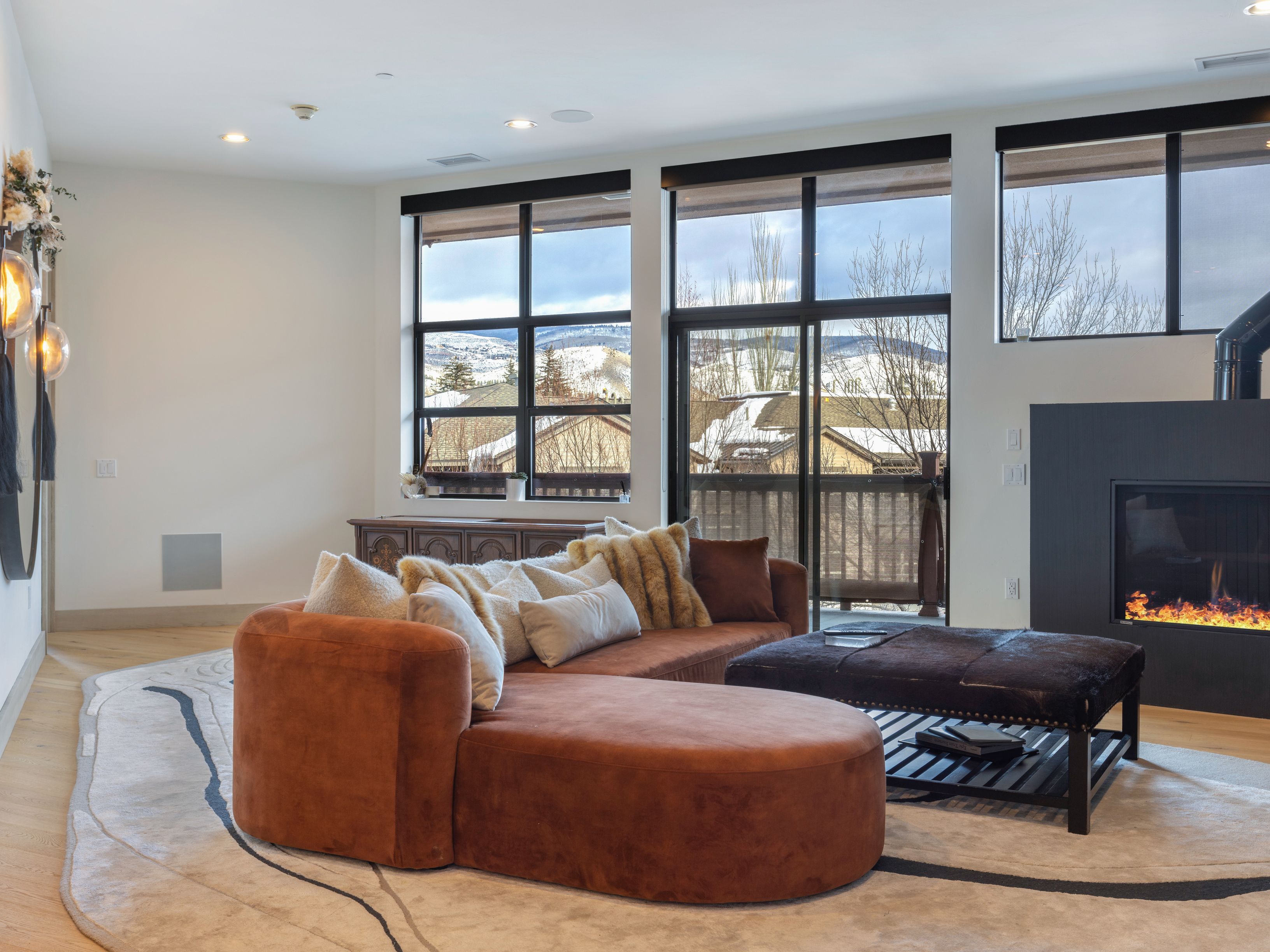This inviting living room features a stylish brown curved sofa adorned with plush pillows, complemented by a dark brown ottoman and a modern fireplace. Large windows offer scenic views, enhancing the room's bright and airy atmosphere. The space is grounded by a contemporary area rug, creating a cozy and sophisticated ambiance.