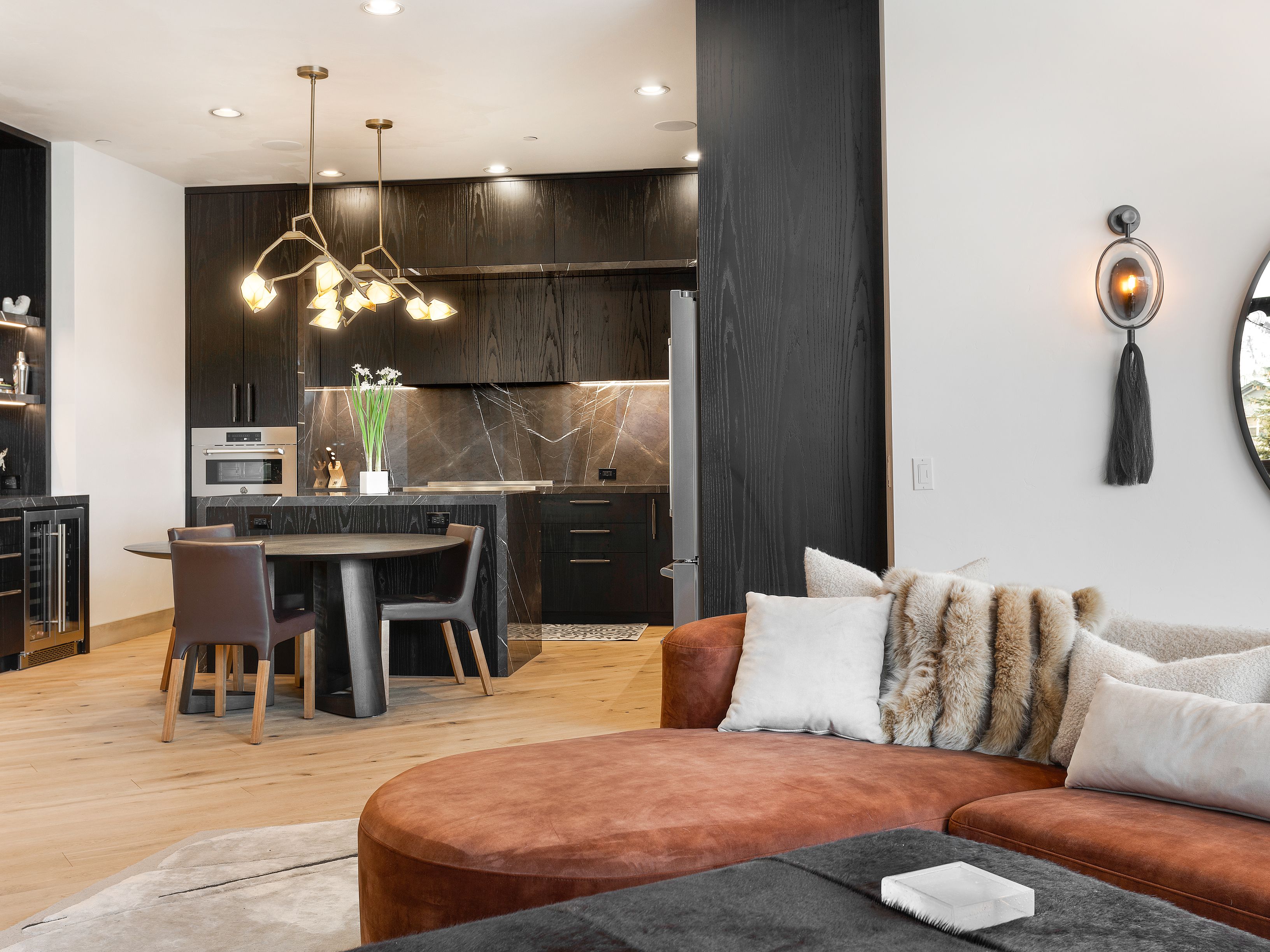 This interior shot showcases a modern living space that seamlessly blends with the kitchen area. The living room features a stylish rust-colored sectional sofa adorned with plush pillows, complemented by a dark gray ottoman. The kitchen boasts dark wood cabinetry, marble countertops, and a unique chandelier, creating a sophisticated and inviting atmosphere.