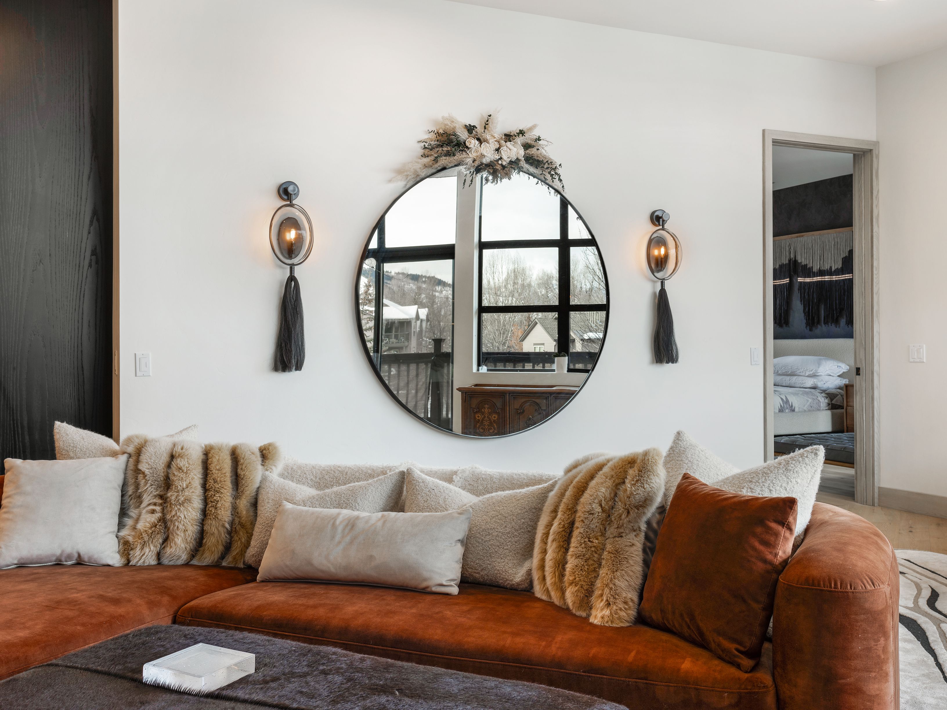 This is an interior shot of a stylish living room featuring a rust-colored sectional sofa adorned with an array of plush pillows in varying textures and tones. A large, round mirror with a black frame hangs on the wall, flanked by sconces with tassel details. The room exudes a cozy and luxurious atmosphere, perfect for relaxation and entertaining.