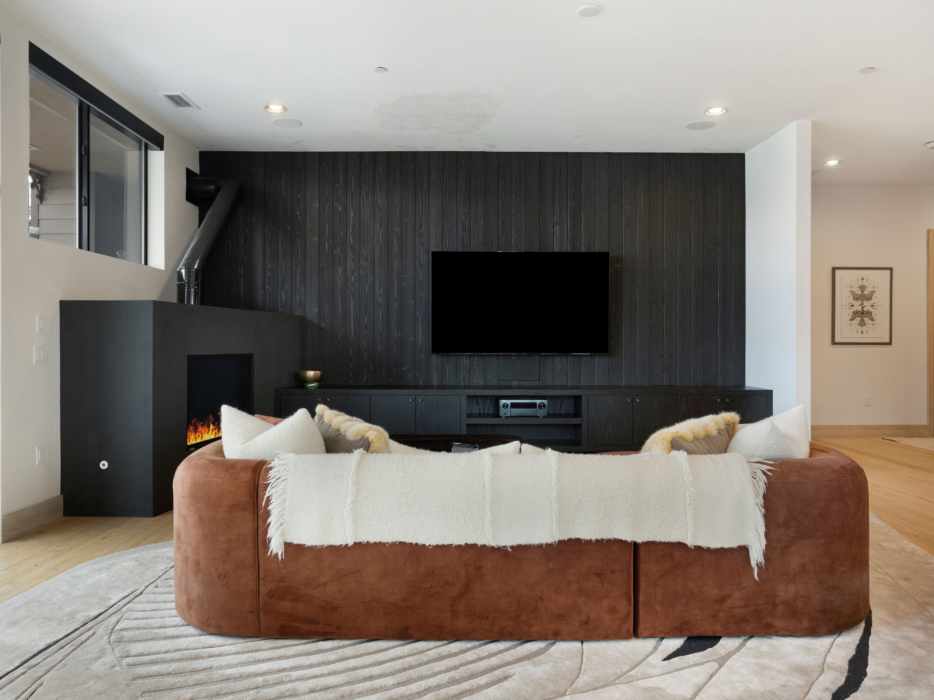 This is an interior shot of a modern living room. The focal point is a large, curved brown sofa with a white throw blanket, positioned on a patterned round rug. Behind the sofa is a dark wood paneled wall with a mounted television and a modern fireplace to the left, creating a cozy and stylish atmosphere.
