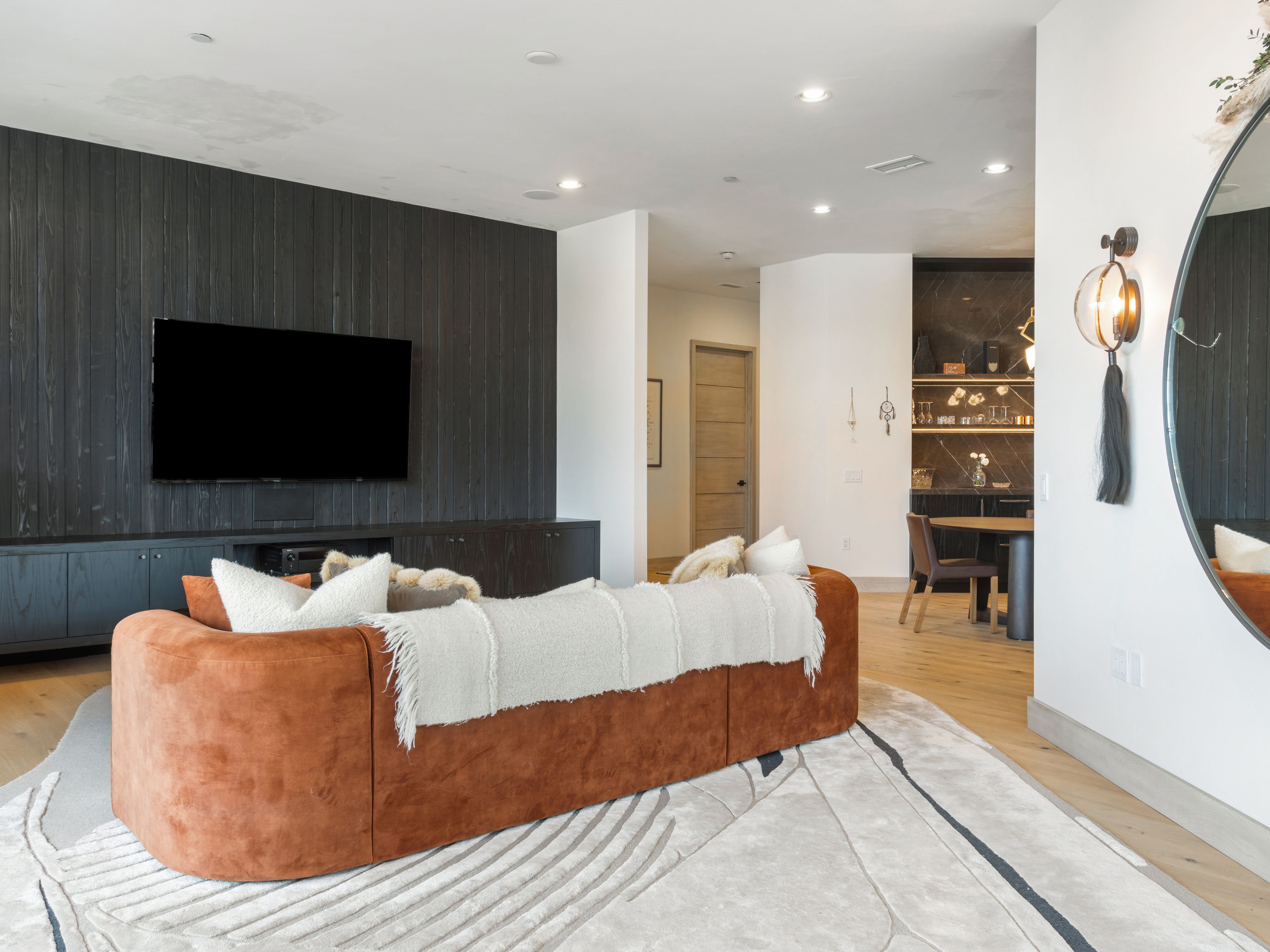 This is an interior shot of a modern living room featuring a large, rust-colored sofa with a white throw blanket. The room has a dark wood accent wall with a mounted television, and a stylish rug covers the hardwood floor. The space is well-lit and appears to be part of an open-concept design, with a dining area visible in the background.