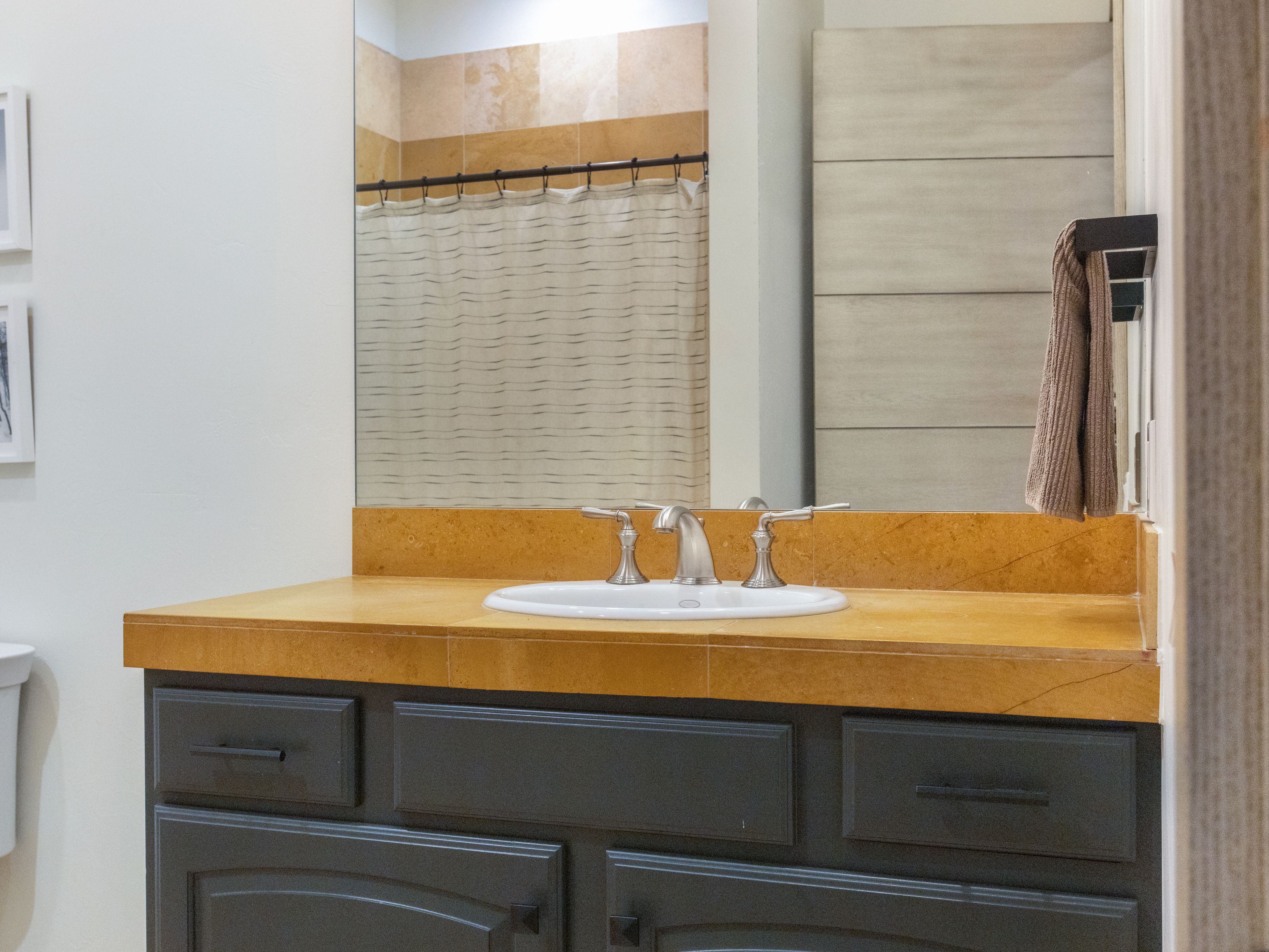 This is a bathroom featuring a vanity with a dark gray cabinet and a light brown countertop. A white sink is centered on the countertop, with a silver faucet. A mirror reflects a shower curtain and a towel rack is visible on the right side of the mirror.