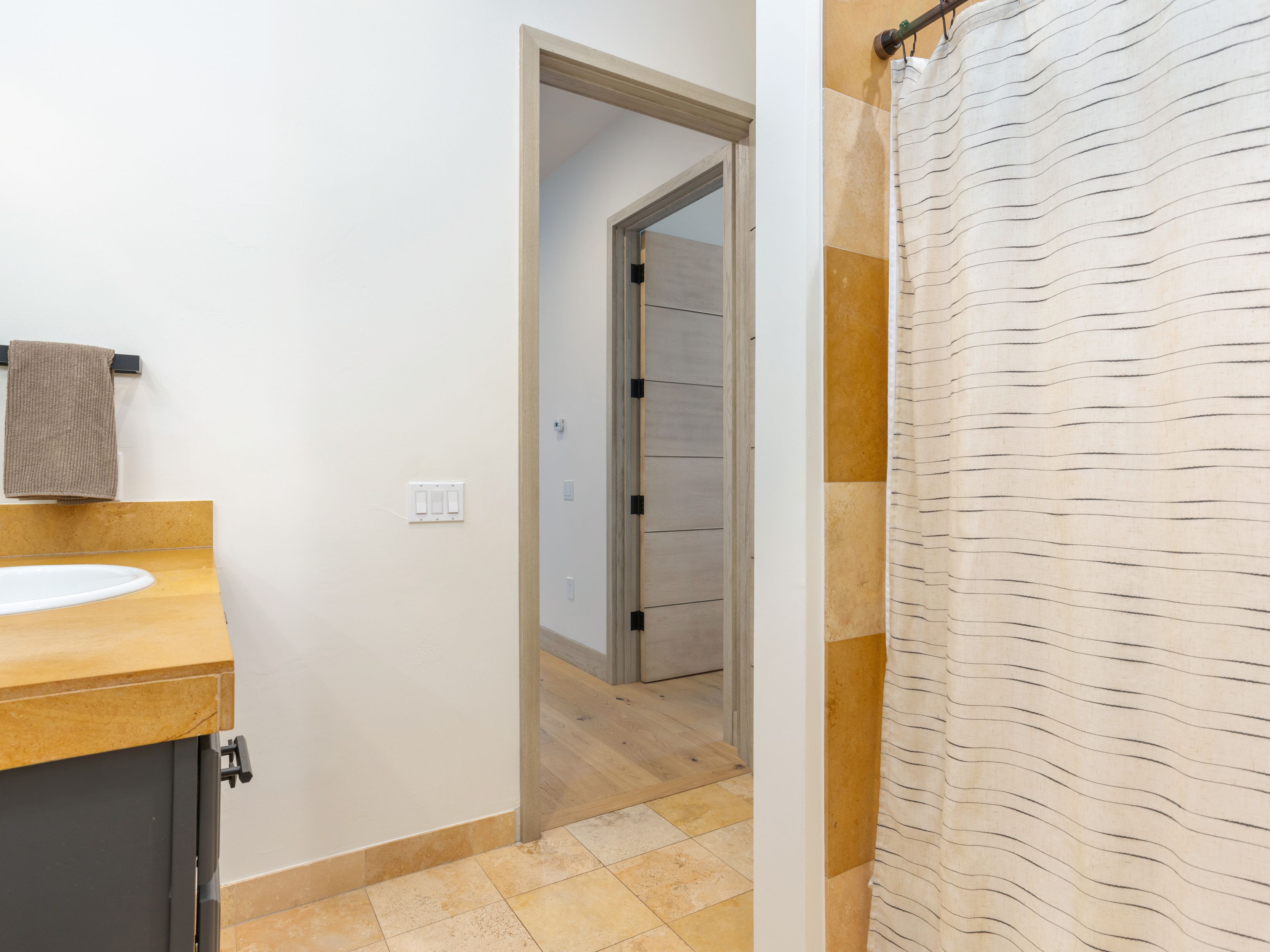 This is a bathroom featuring a vanity with a light-colored countertop and dark cabinetry. The walls are a neutral color, and the flooring is tiled. A doorway leads to another room, and a shower with a patterned curtain is visible on the right. The overall impression is clean and functional.