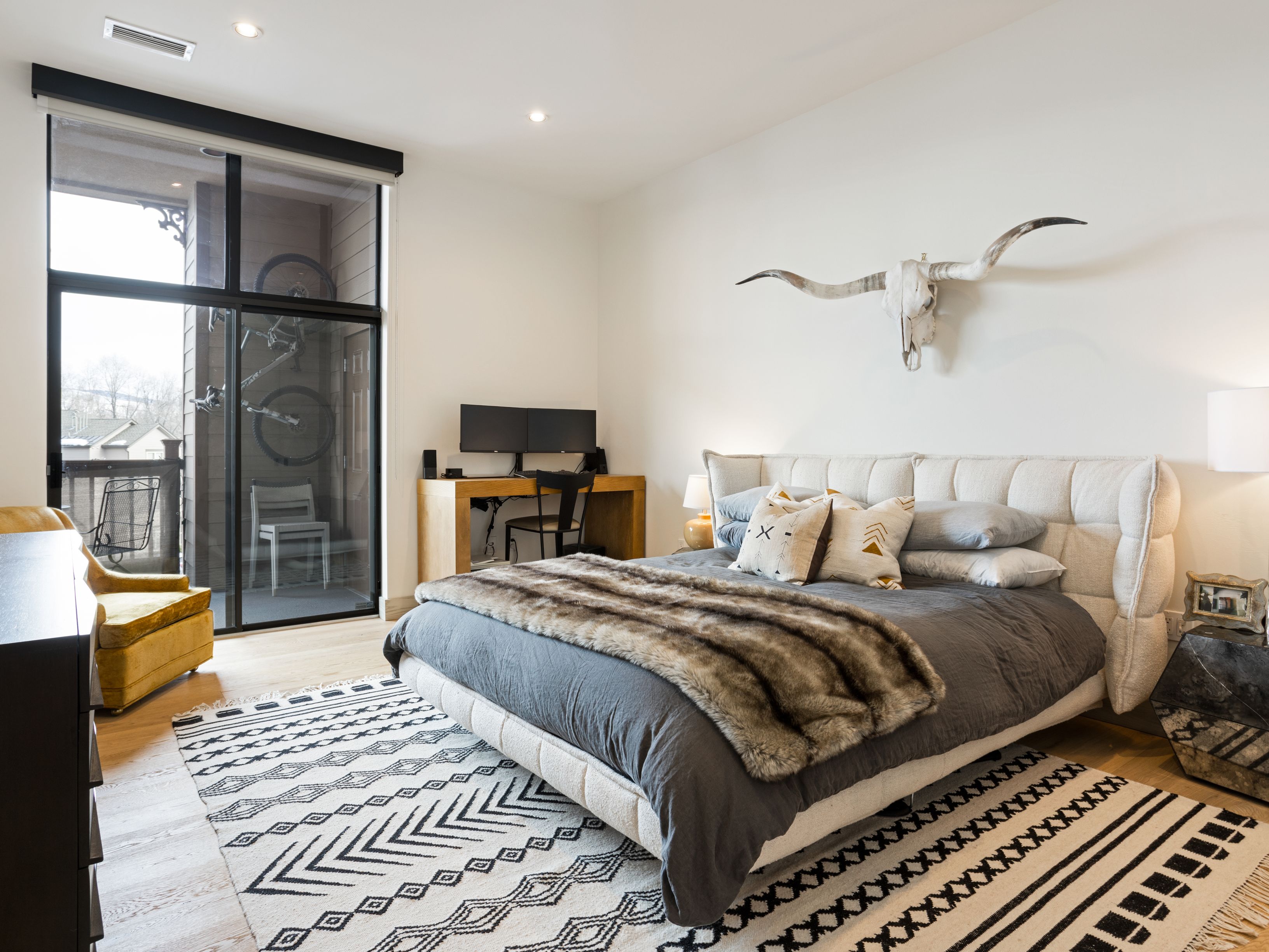 This is a well-lit primary bedroom featuring a large bed with a tufted headboard, a gray duvet, and a faux fur throw. A desk with a computer setup is positioned against one wall, while a decorative steer skull adorns another. The room opens to a balcony with a bicycle rack, and a patterned rug adds visual interest to the hardwood floor.