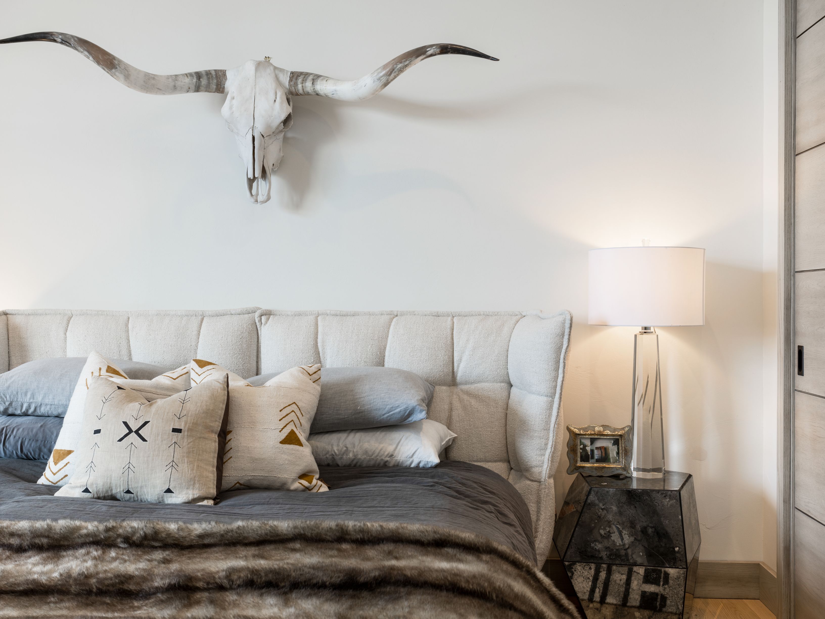 This is a well-decorated primary bedroom featuring a large bed with a tufted headboard, decorative pillows, and a faux fur throw. A longhorn skull is mounted above the bed, adding a rustic touch. A modern lamp sits on a mirrored nightstand, and a closet is visible on the right, creating a cozy and stylish atmosphere.
